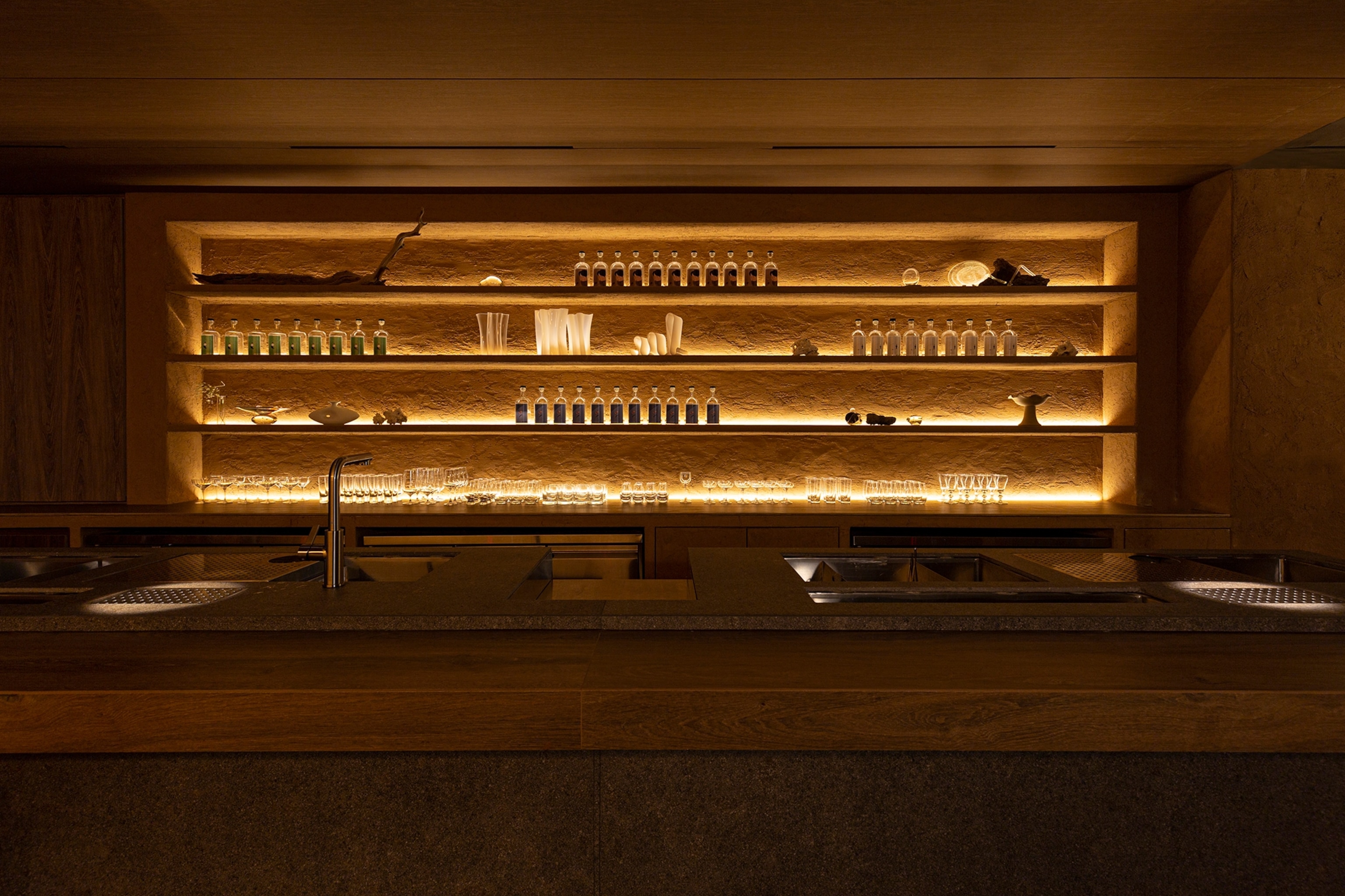 The interior of a moody bar with dimmed lighting coming from behind glass shelves.