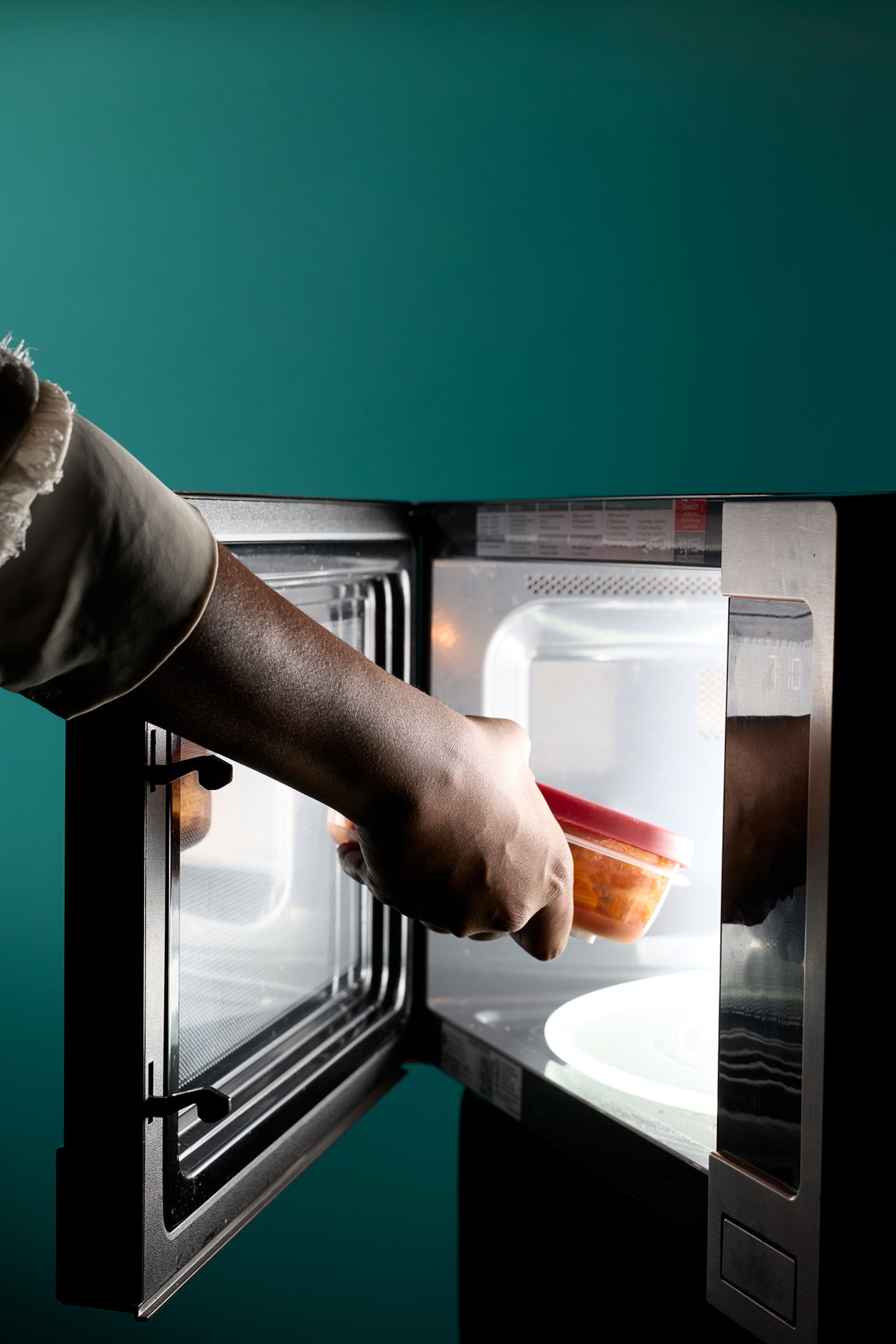 A hand with dark skin tone places a container in a microwave with glowing light.
