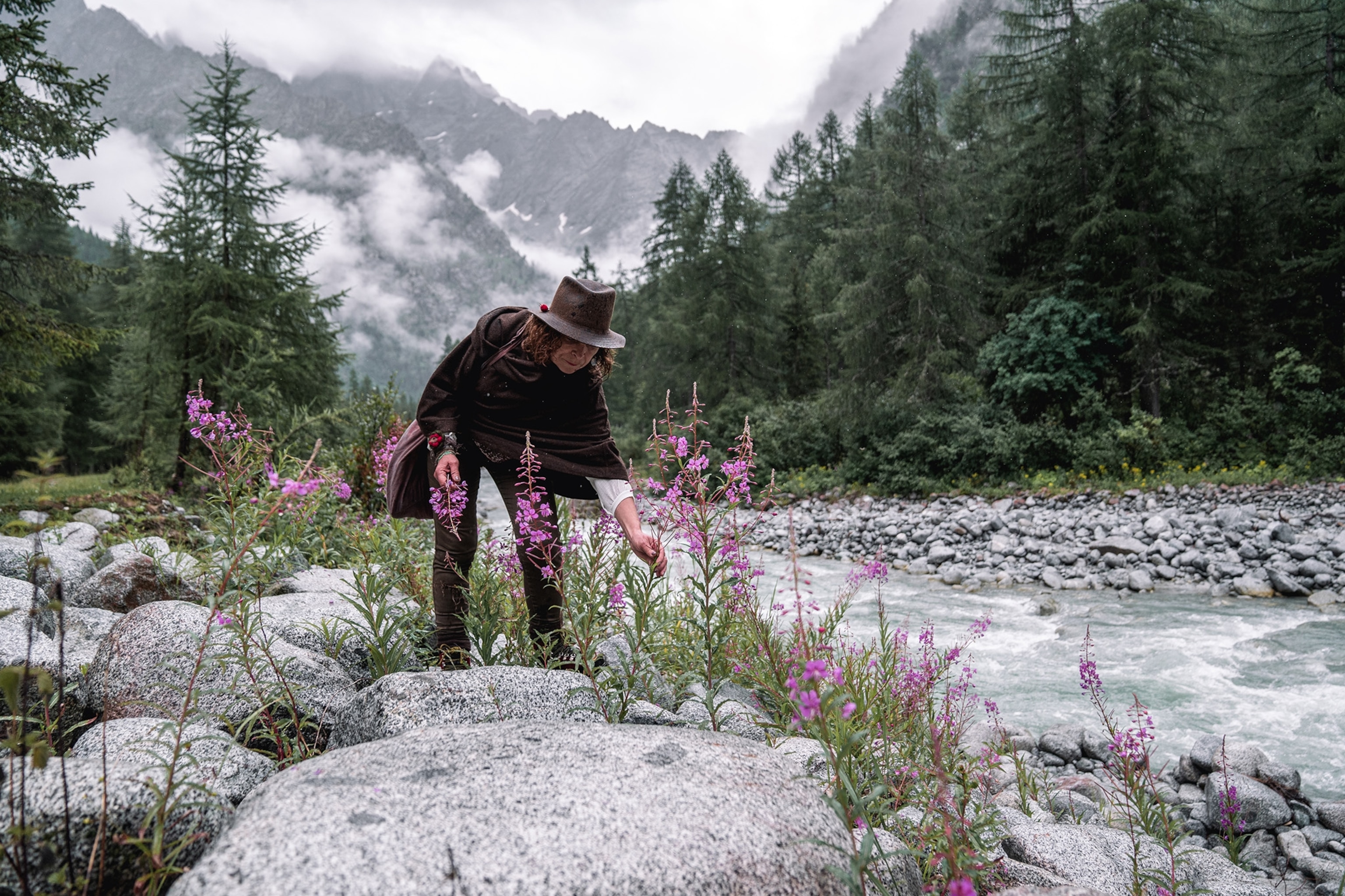 A woman wearing cape and felt hat crouching down on the side of a mountain river to pick a flower.
