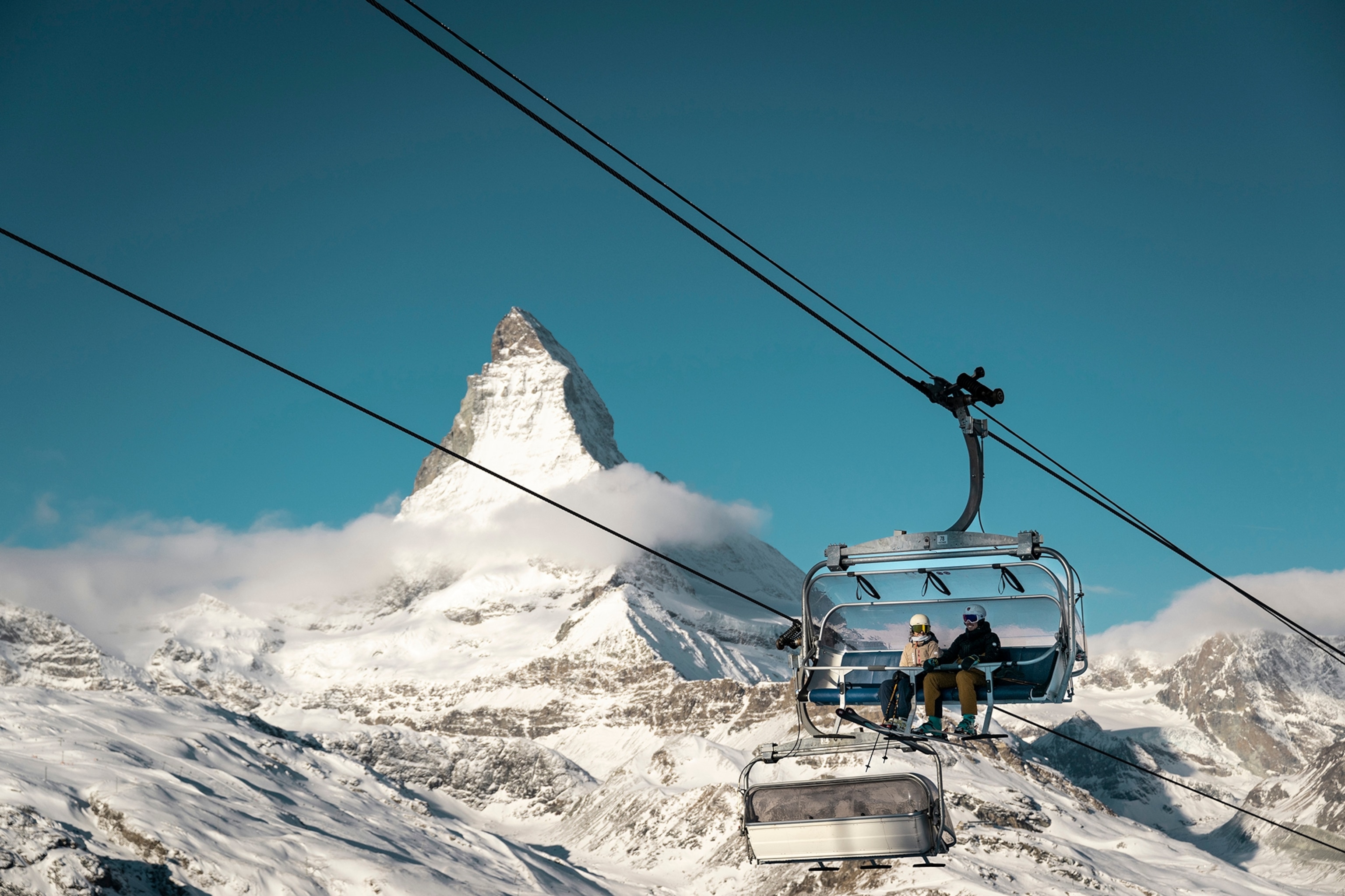 Two skiers sitting in a chair lift with a snowy mountain scape in the background.