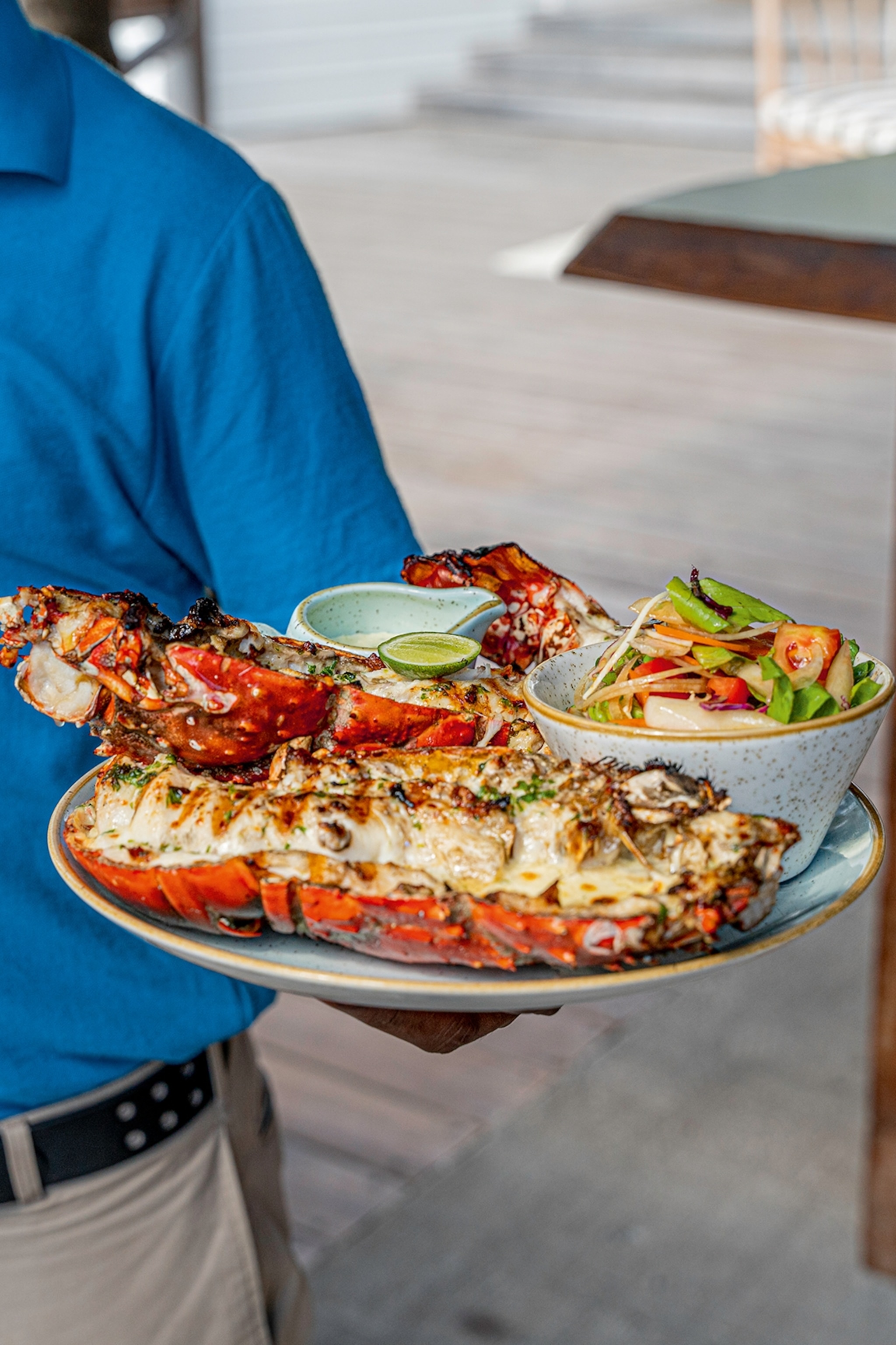 A close up of a waiter holding up a dish of char-grilled lobster served with a tropical side salad, lime wedge and sauce.