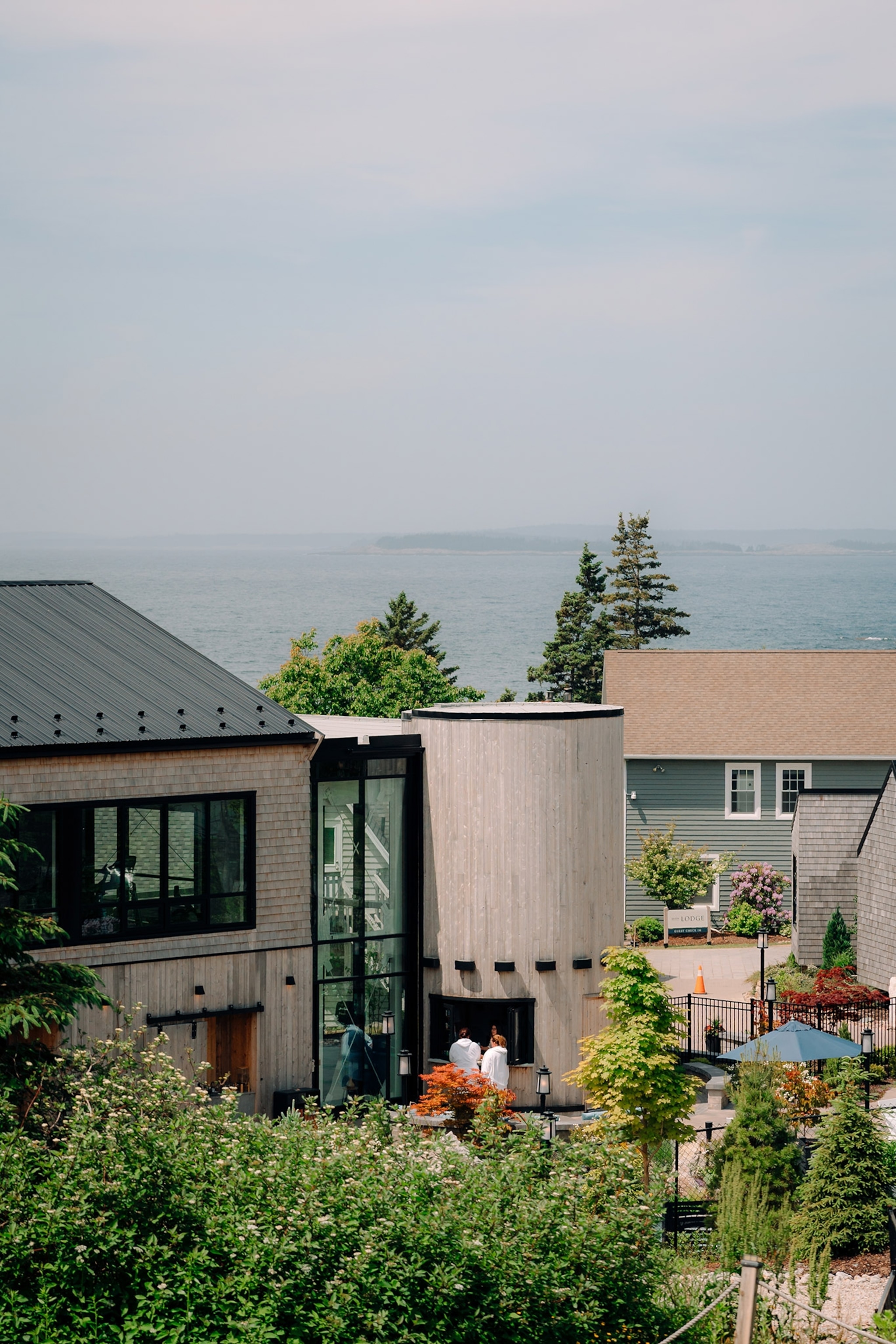Exterior of spa with modern architecture, visitors in white bath robes entering cylindrical buliding, body of water in the distance