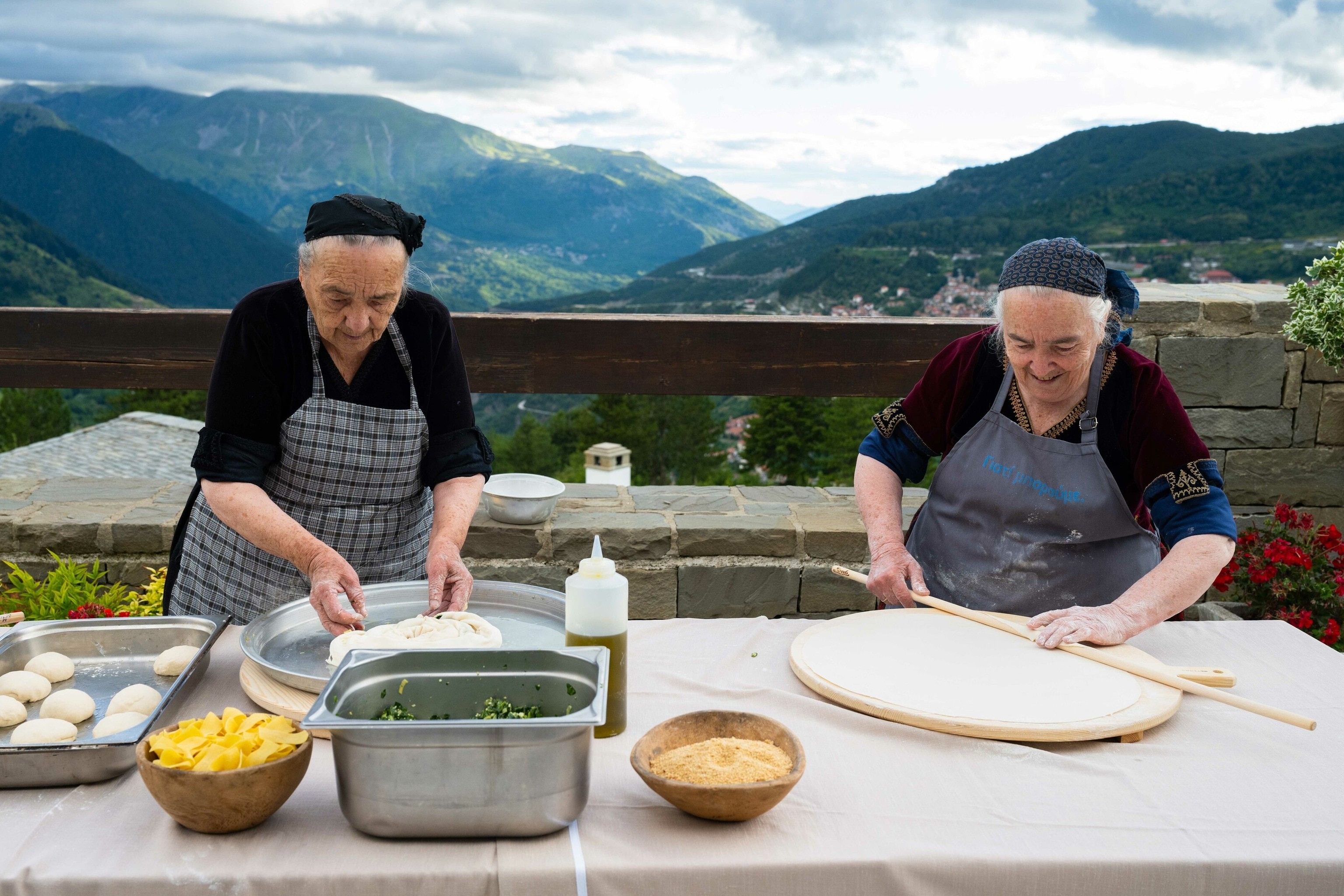 Two women make 'petoura' on large circular wooden boards. One is shaping dough with her hands, the other smooths it out with a long, flat stick.