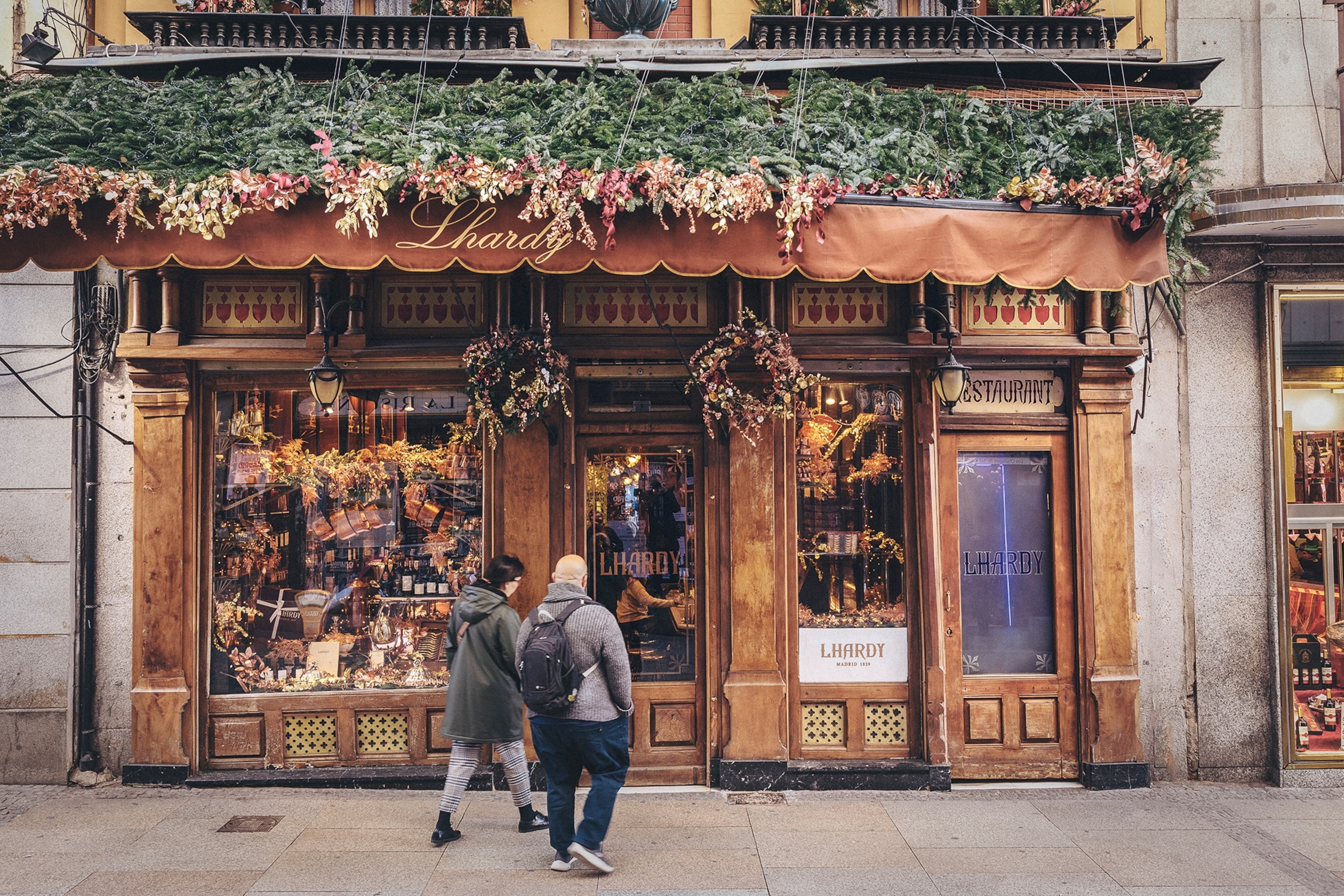 A street-side restaurant with a weathered wooden facade and flowers draped over the top.