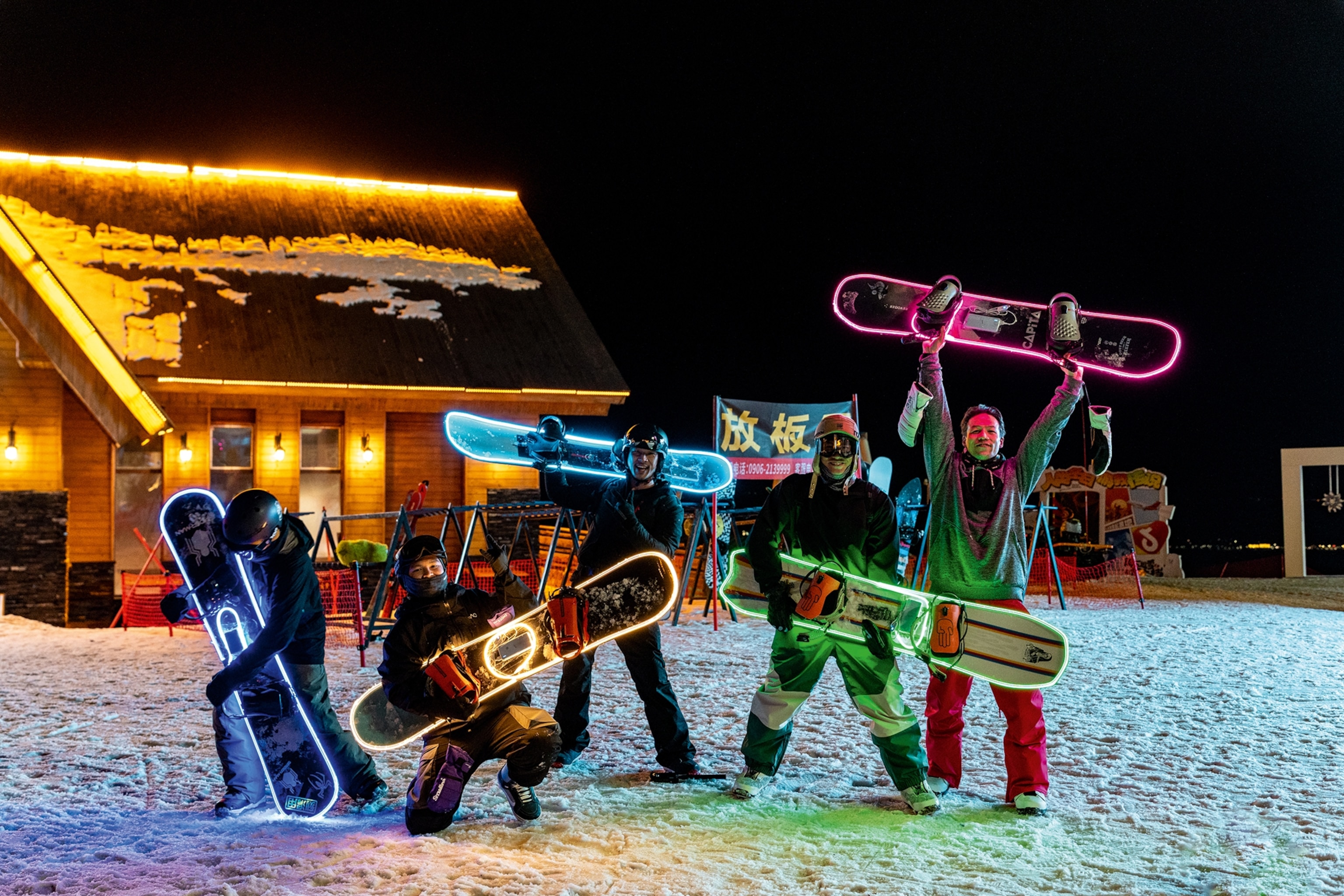 A group of snowboarders holding up their neon-lit boards outside a chalet at night.
