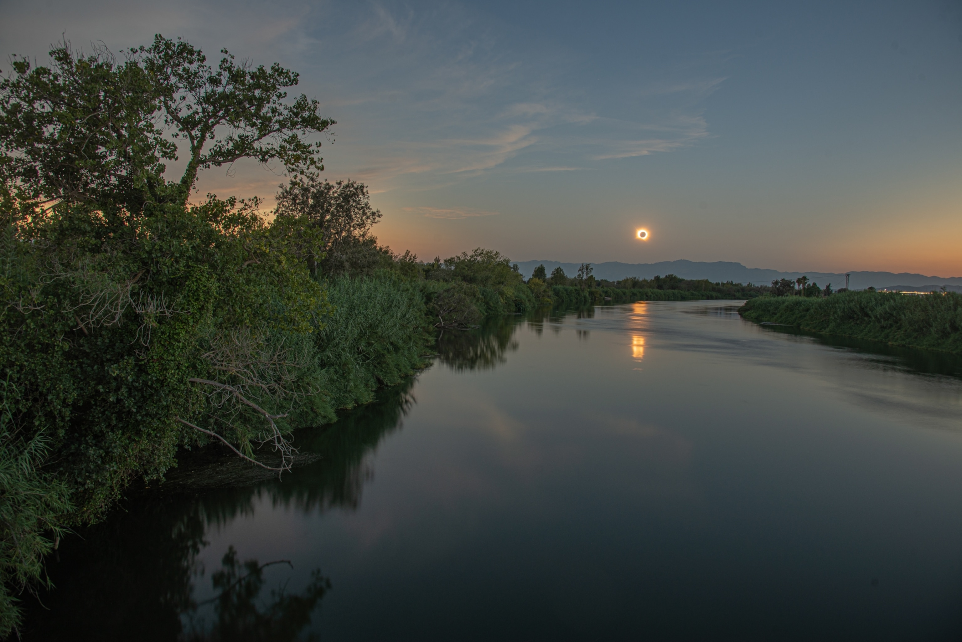 A tranquil river at dusk reflects the orange eclipse, framed by leafy reeds and trees, with distant mountains beneath a fading blue sky.