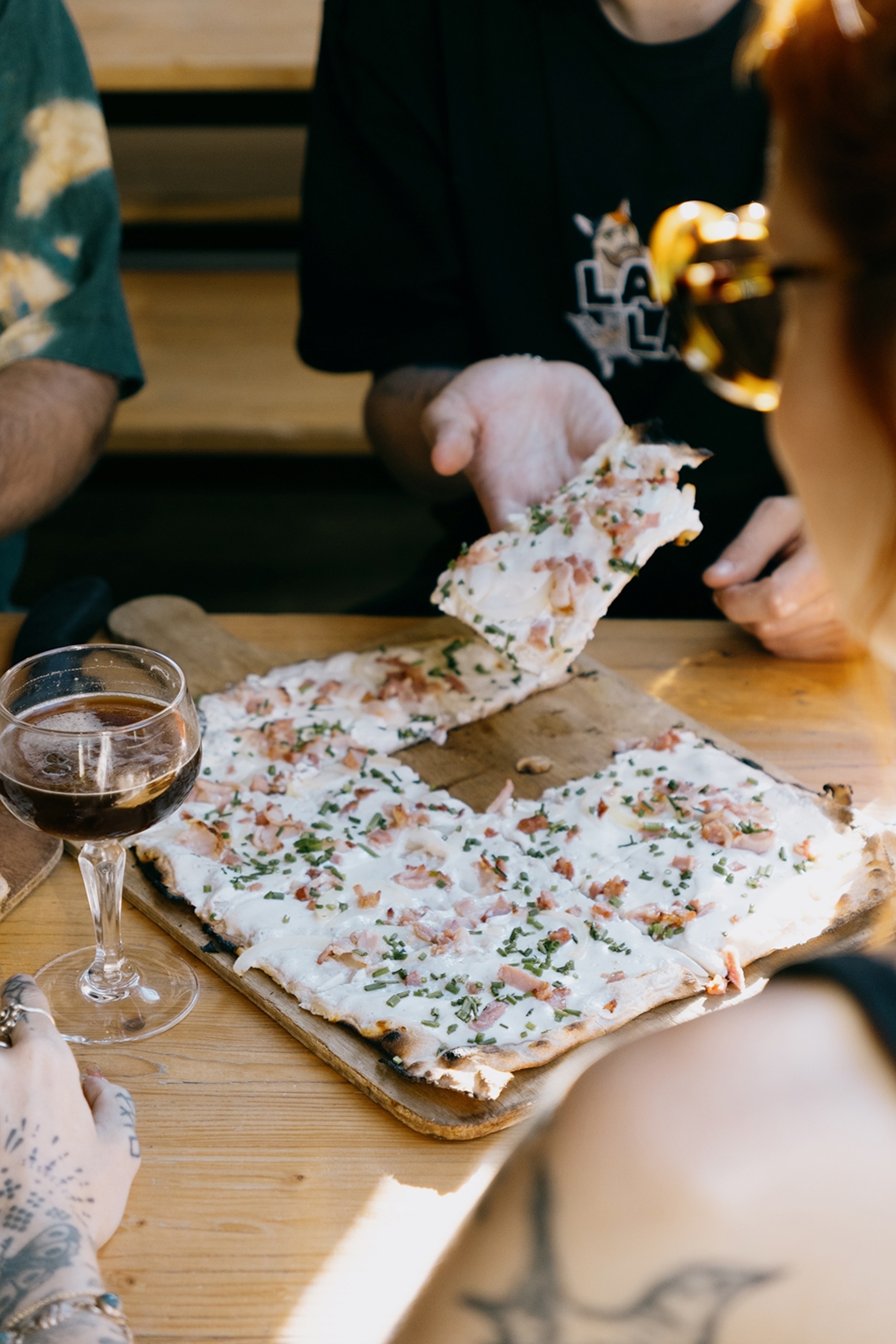A thin tart with cream cheese and ham bits on a rustic wooden board as diners take square slices.