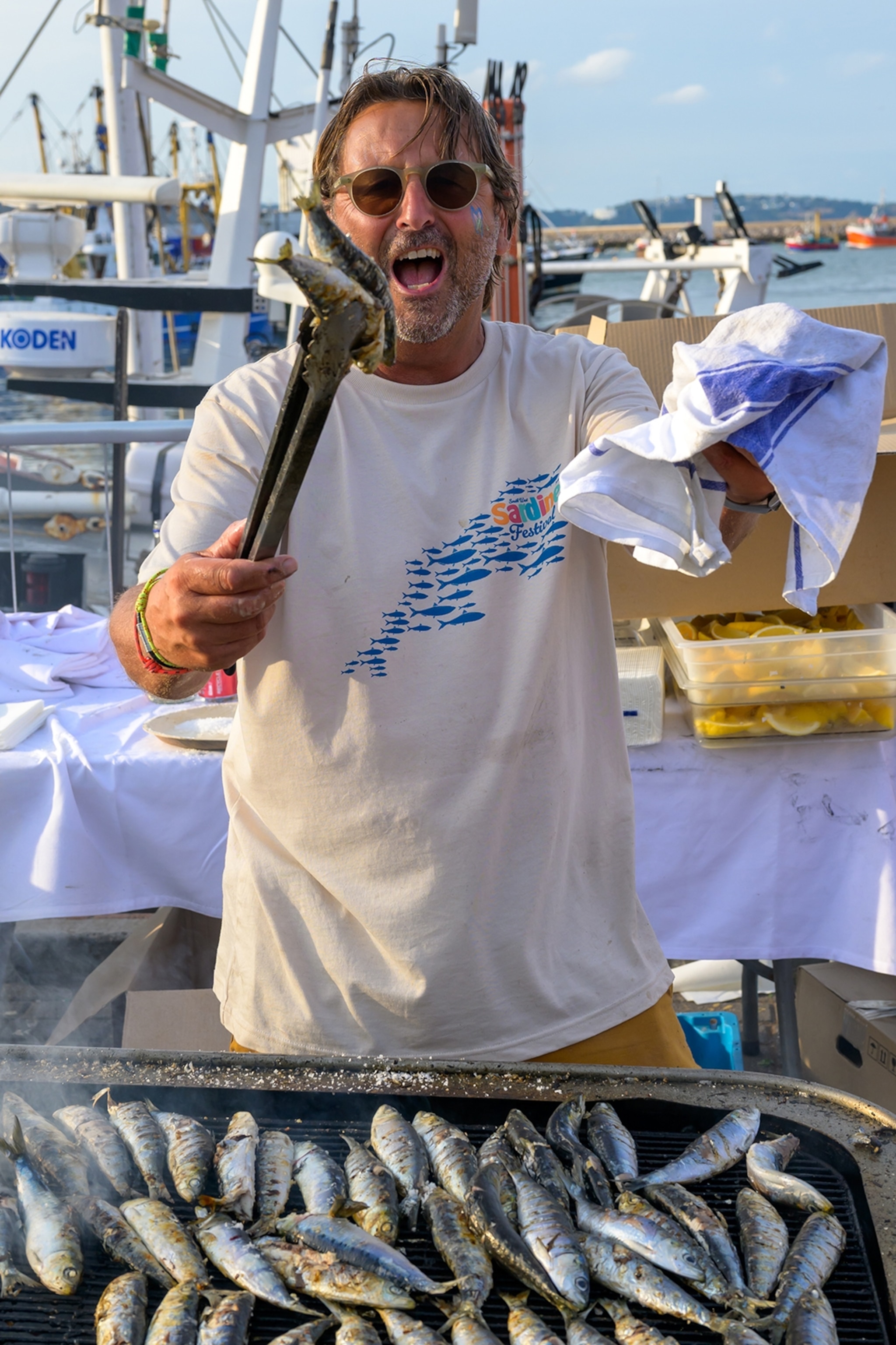 A chef behind a barbecue holding up sardines from the grill.