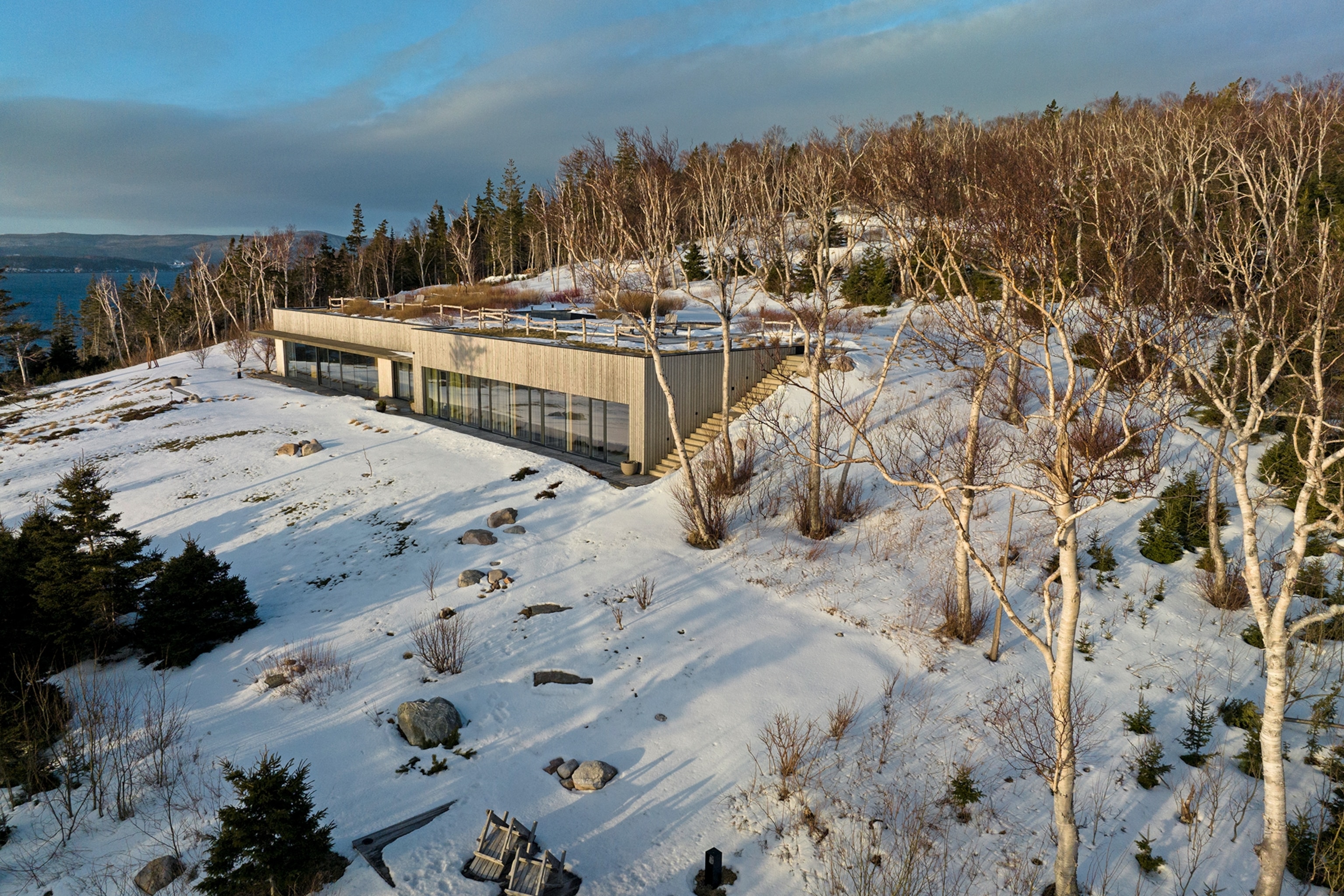An side-view shot of a modernist concrete villa set into the side of a snowed-in hill with a bare forest in the back.