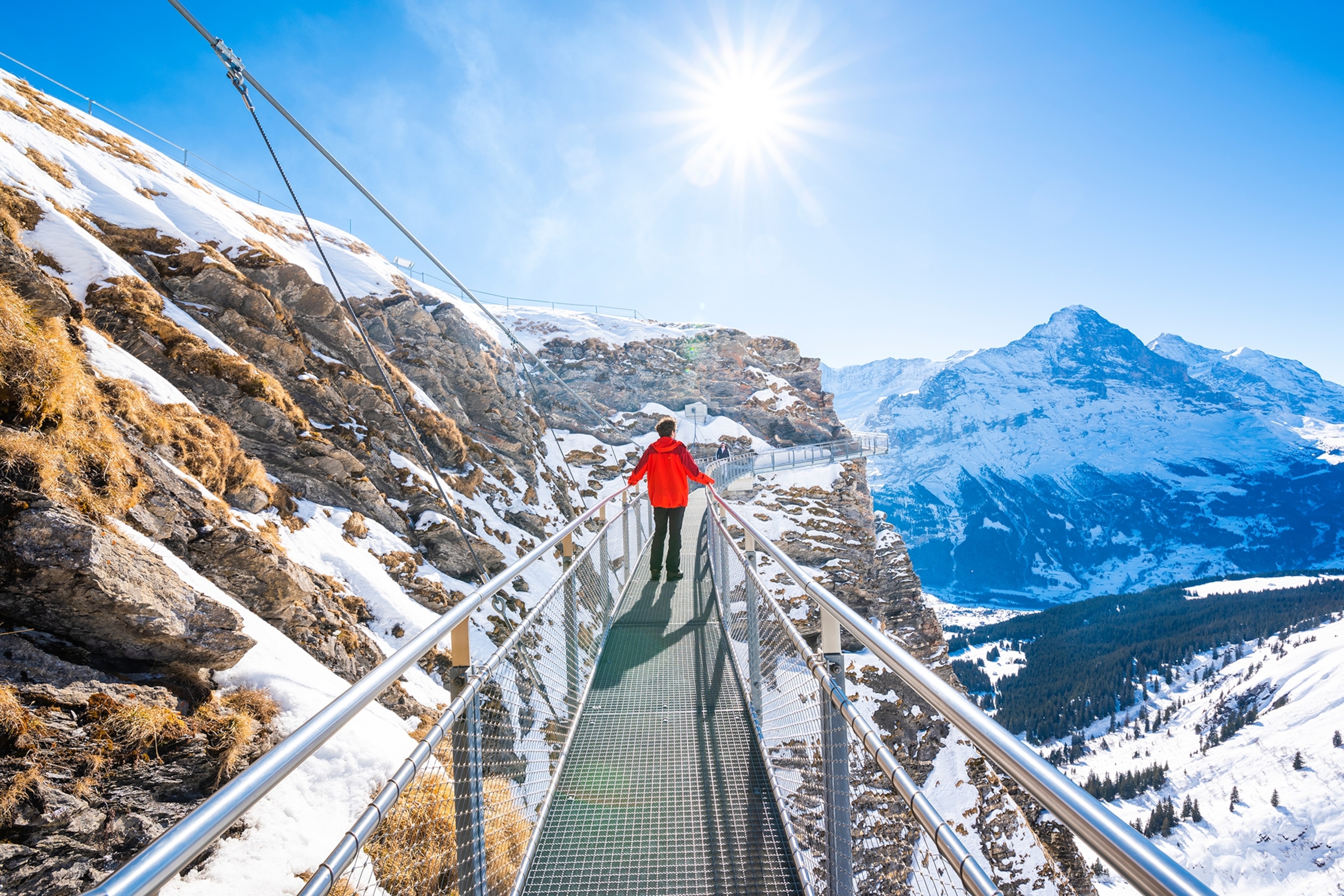 A man in a red jacket stands on a walkway suspended high amongst the mountains in Switzerland.