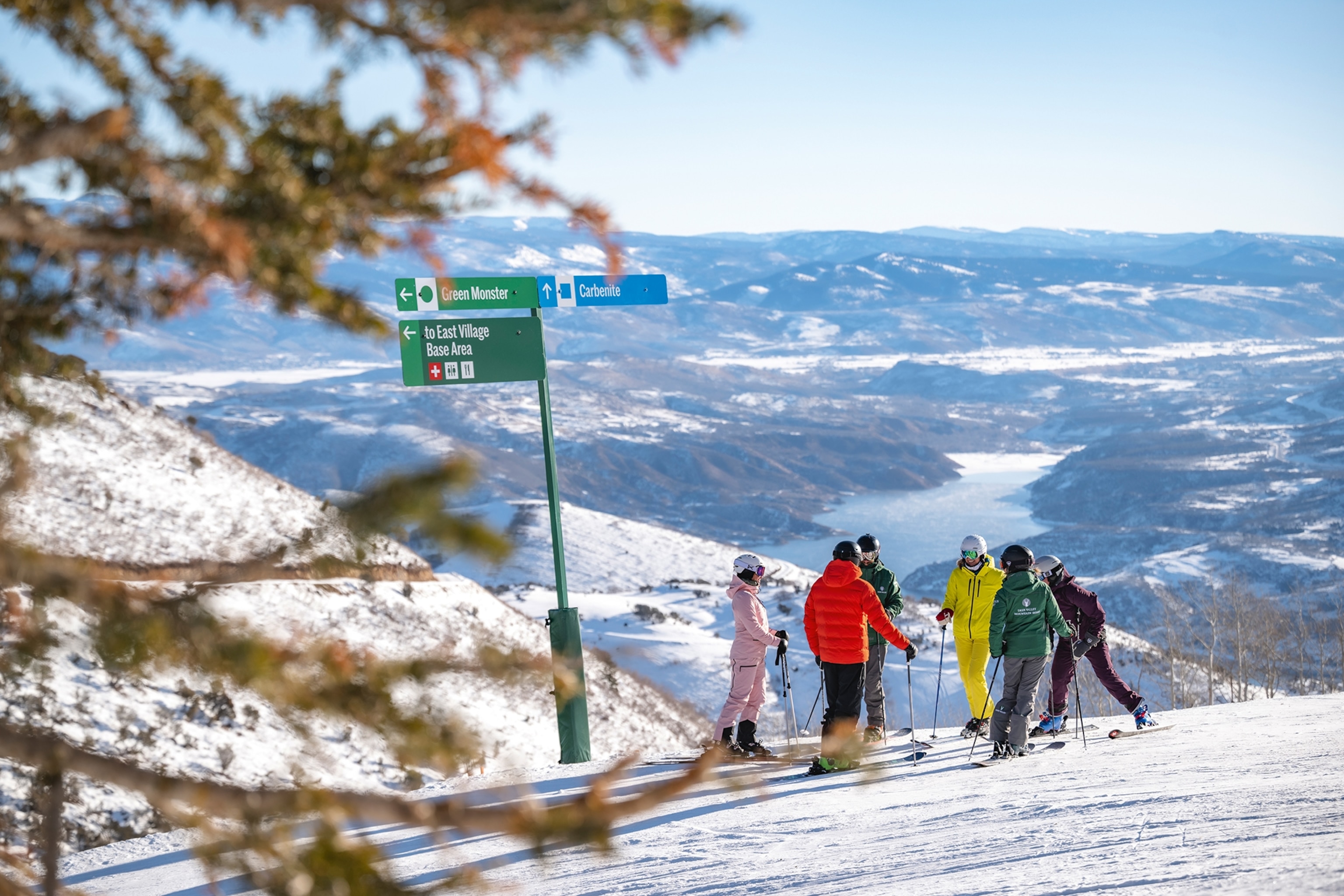 A snowy piste view into a mountain valley as a group of skiers stands on the edge.