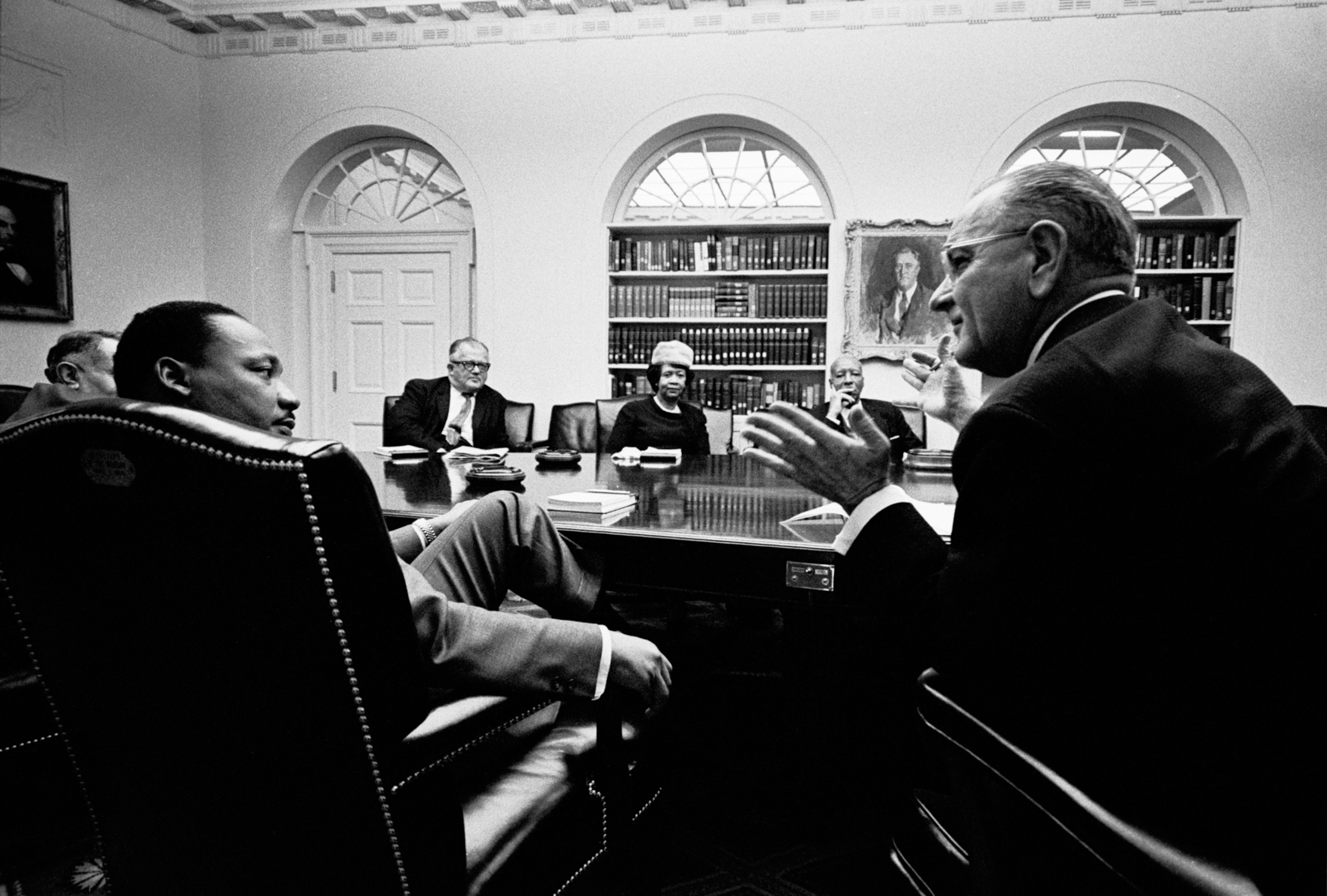 A group of sitting around a table in a formal meeting room with bookshelves and arched windows.