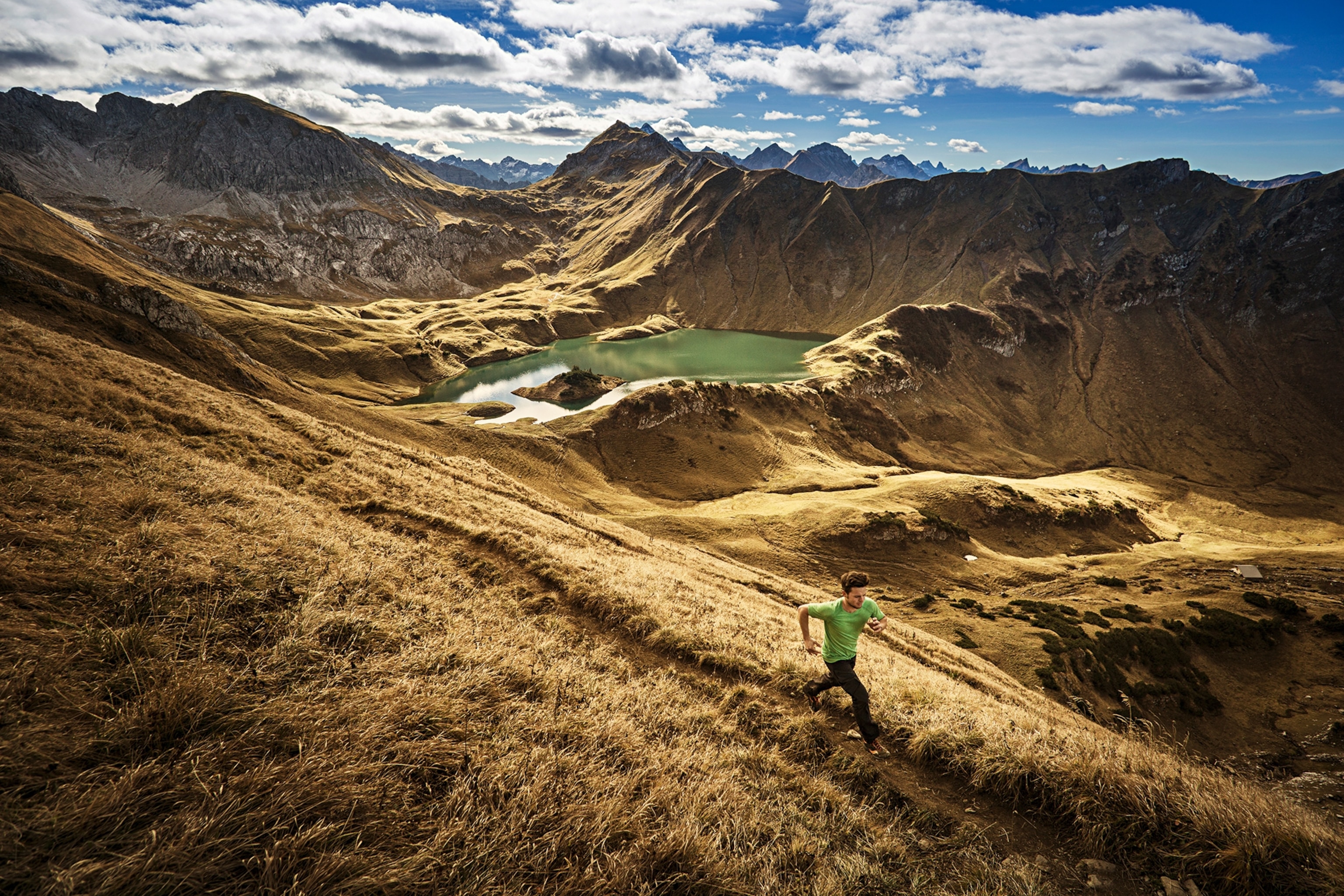 A young, male runner taking a mountain-side path with a range and lake in the background.
