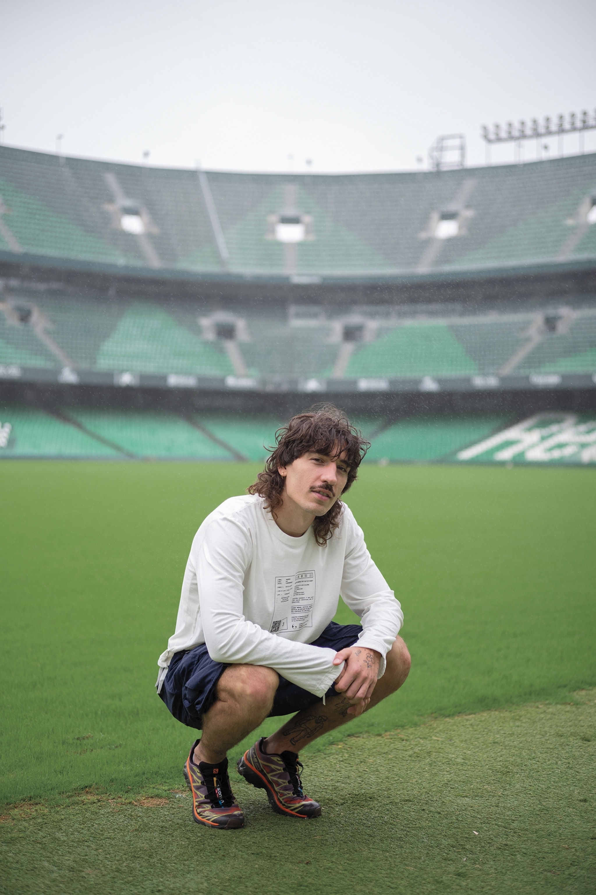 A man squats and looks at the camera for a portrait. He is on an empty stadium field. There are stadium seats in the background.