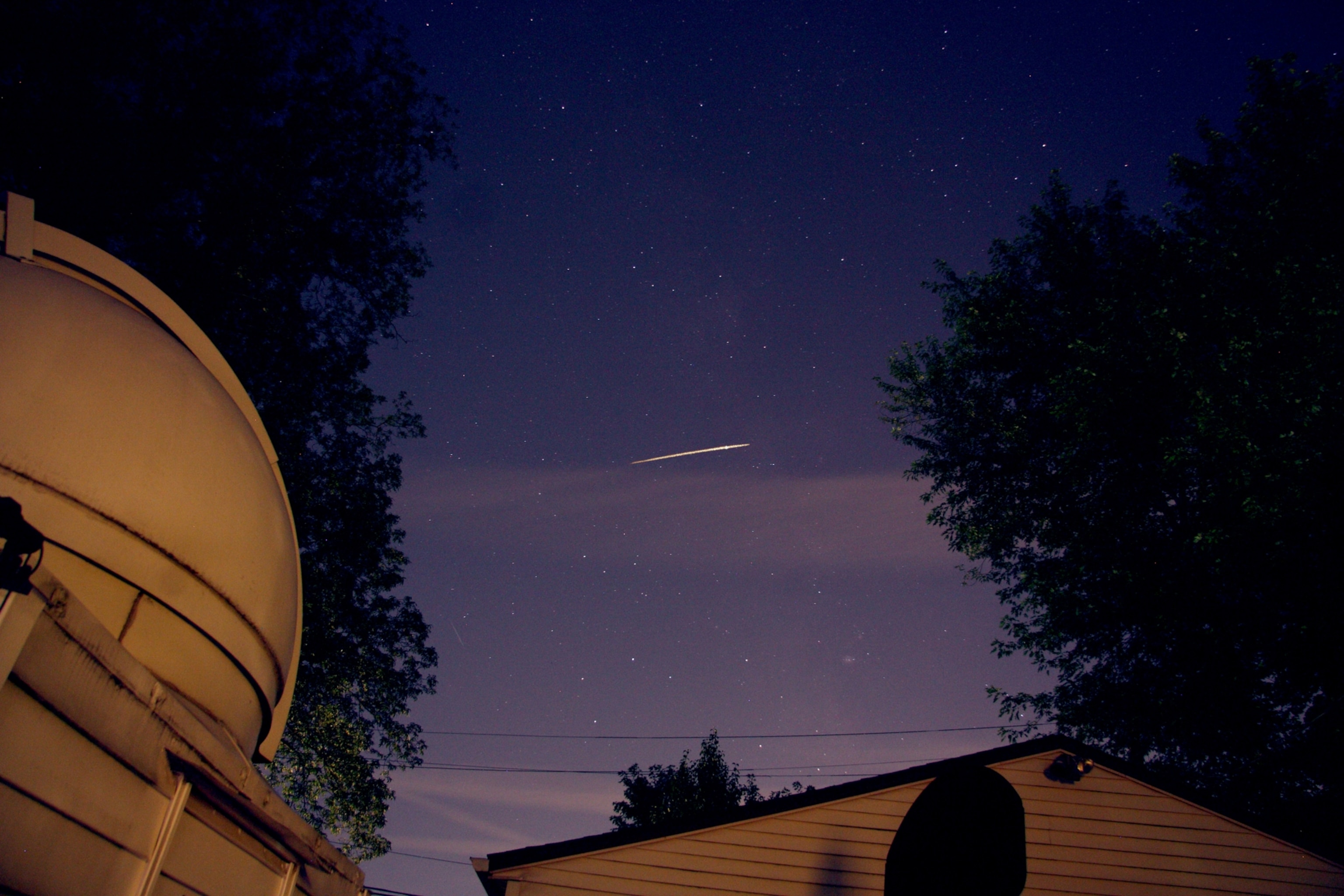 Night sky with numerous stars over rooftops, featuring a bright meteor streak across the center