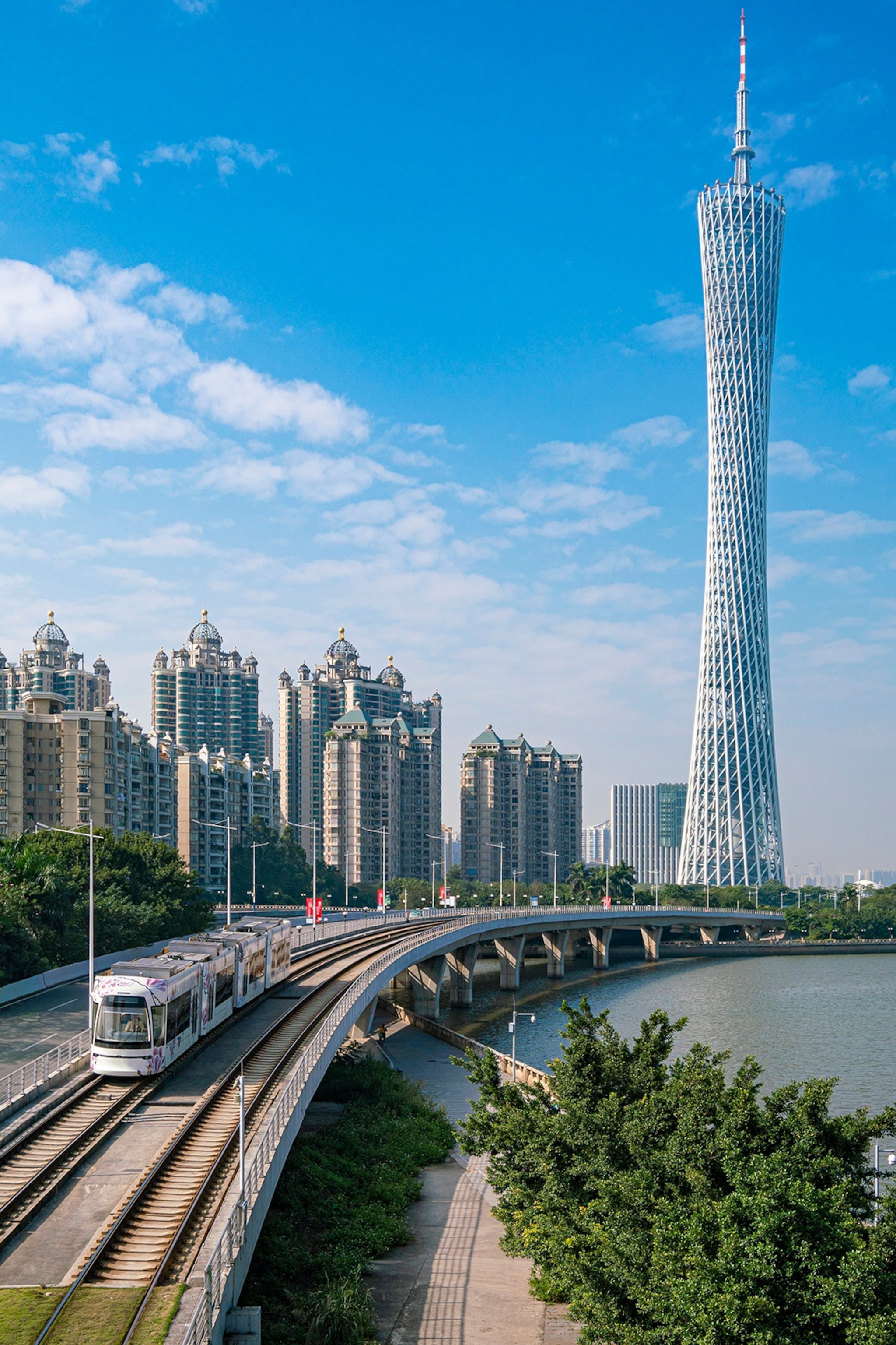 A train running along the river with a metropolis bordering on the other side as a skyscraper reaches into the sky.