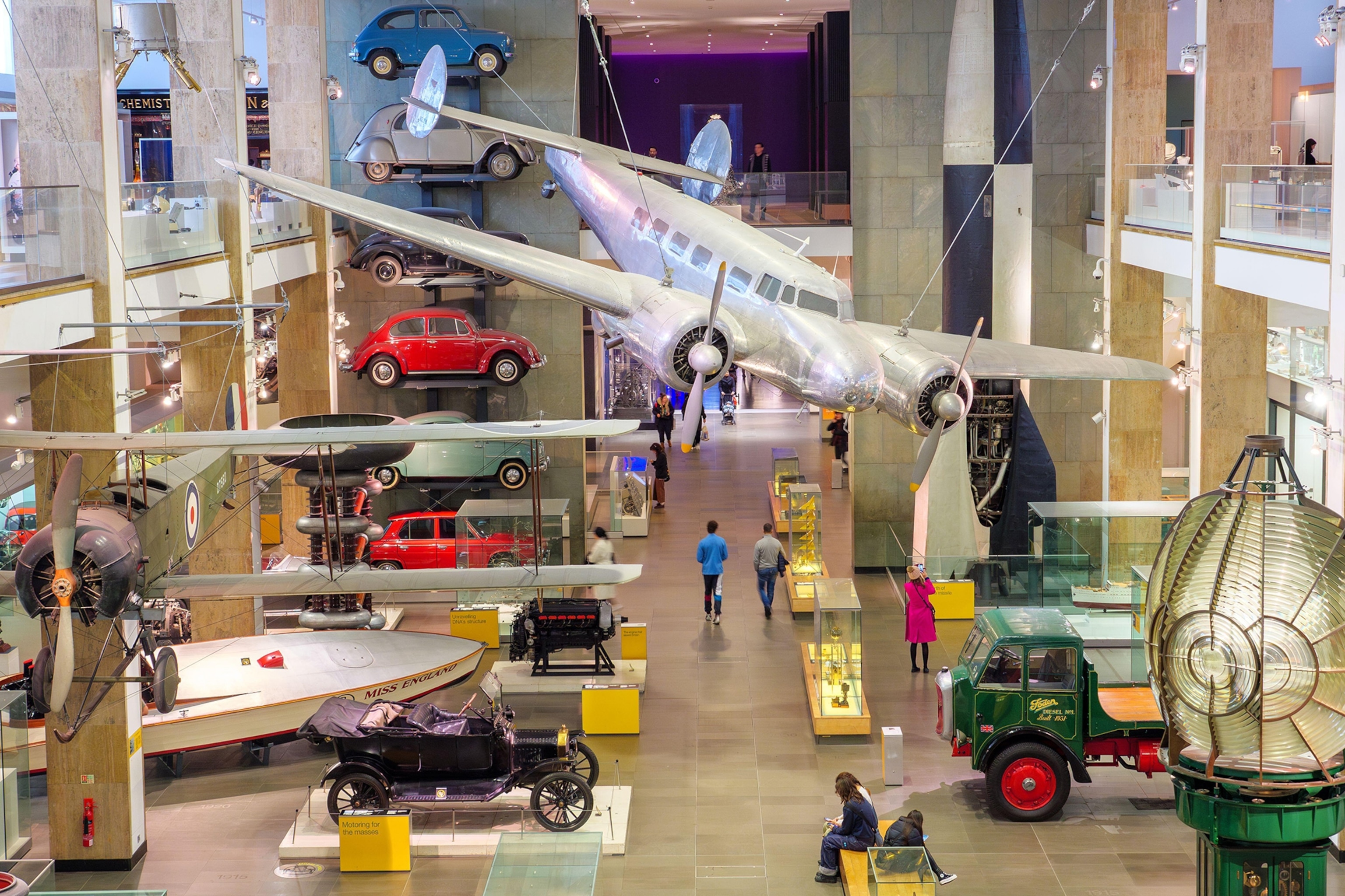 A big museum hall with old cars and other modes of transport exhibited on the walls and grounds as a steel plane is suspended in the air.