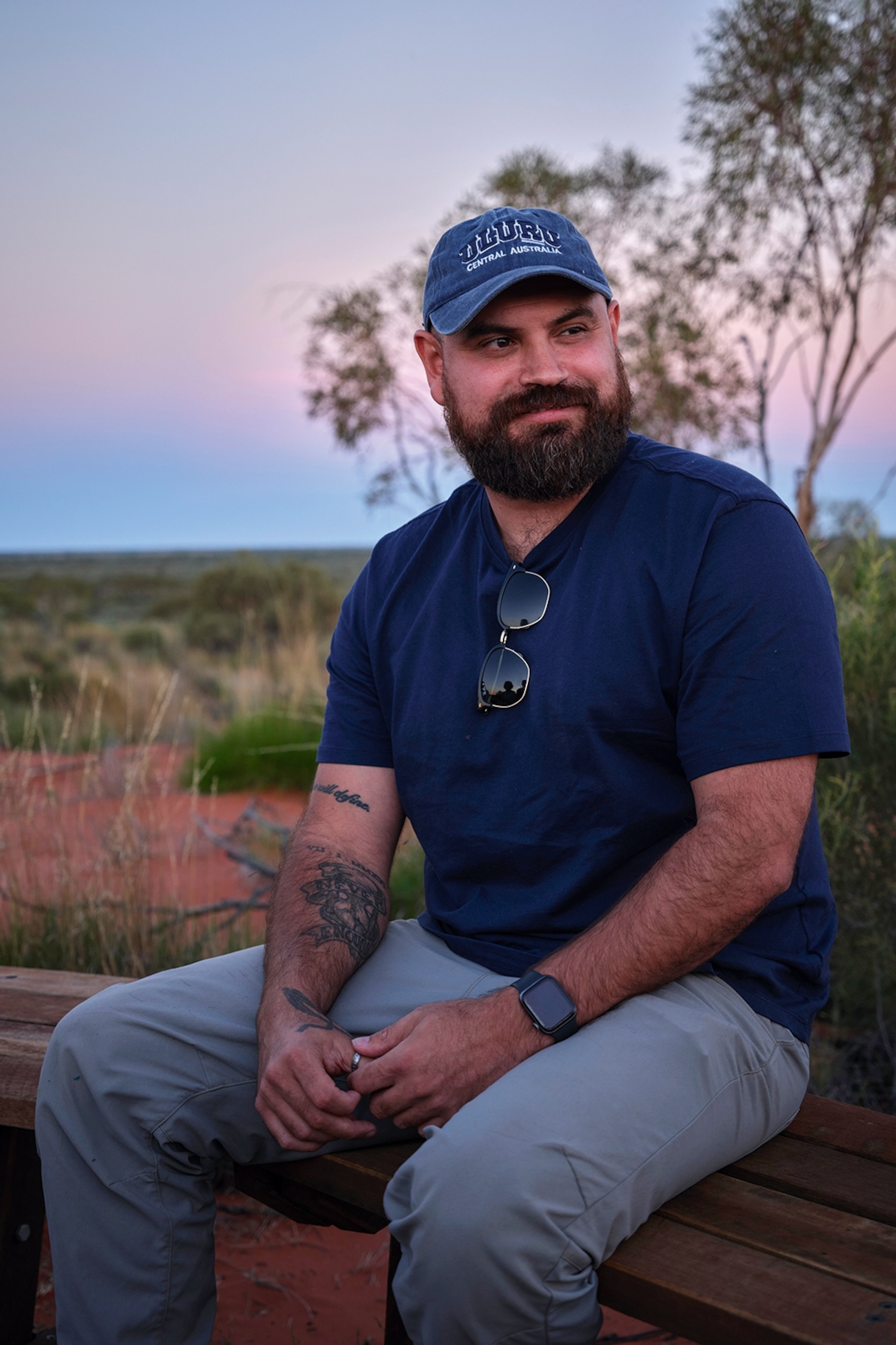 A young man with a moustache sitting on a wooden bench in the Outback.