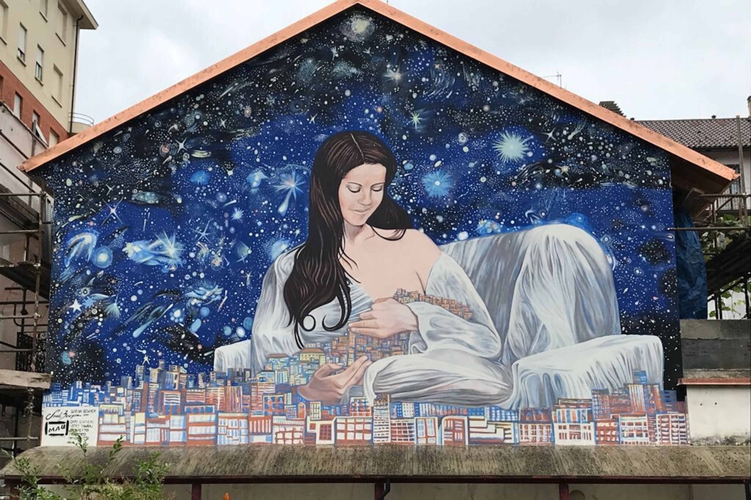 A mural of a woman cradling a city like a baby. She is surrounded by stars, and appears to be breast feeding the city.