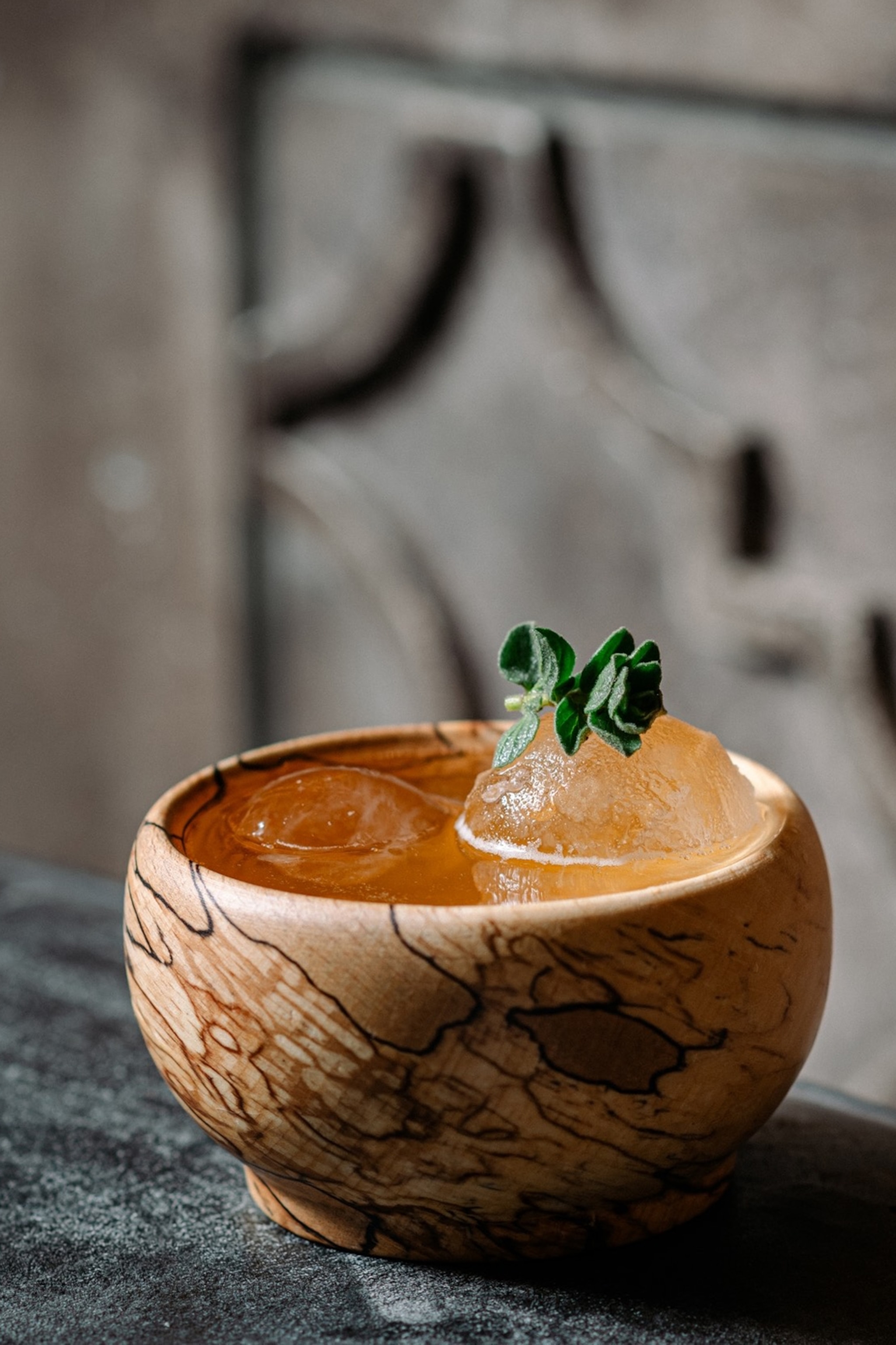 A close-up of a cocktail in a wooden bowl with round ice cubes and fresh oregano on top.