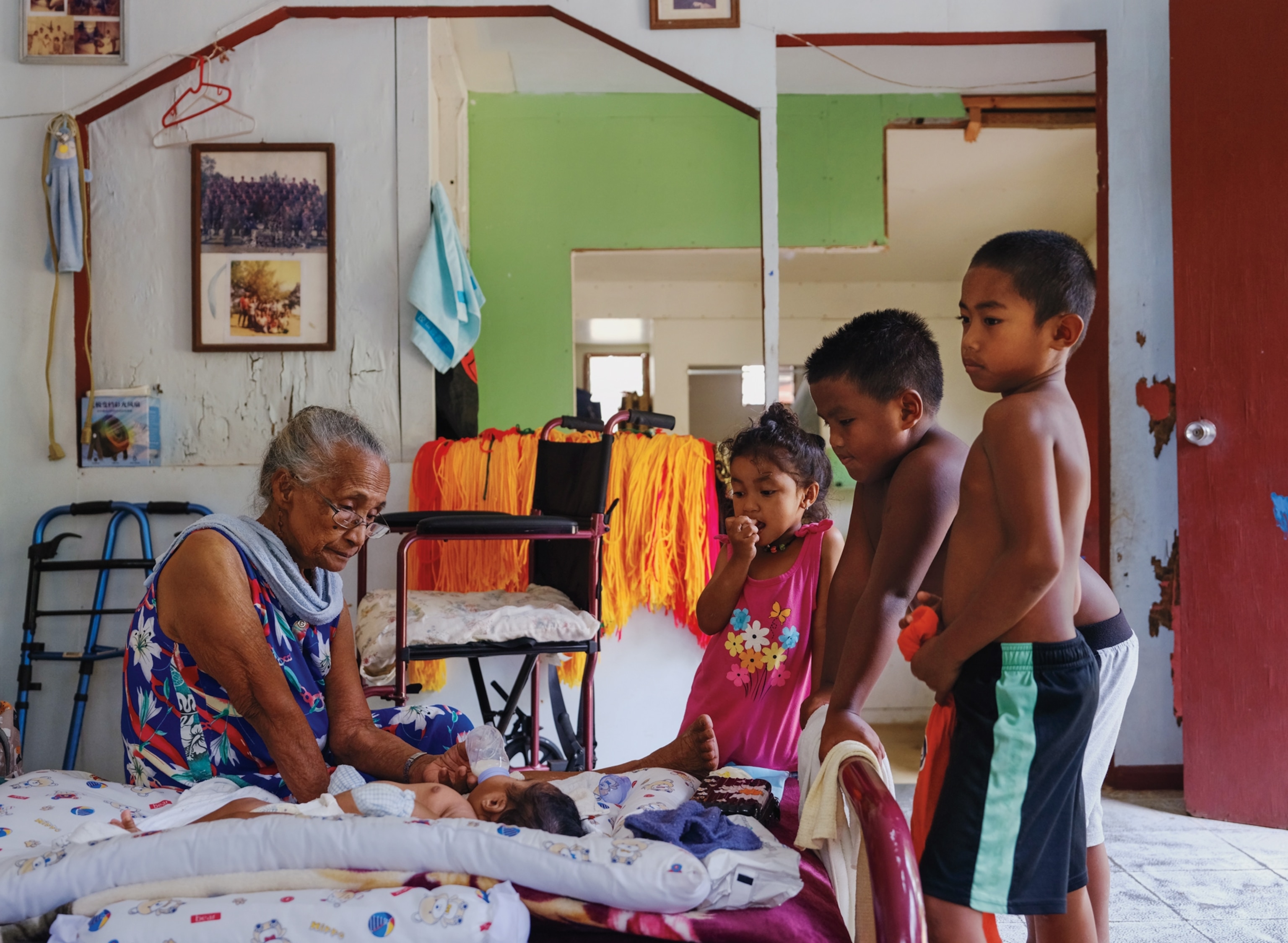 An older woman sits on a bed and holds a bottle while feeding a baby. To the right are three young children watching her. The older woman and young girl wear sleeveless dresses, while the two young boys wear shorts and no shirts.