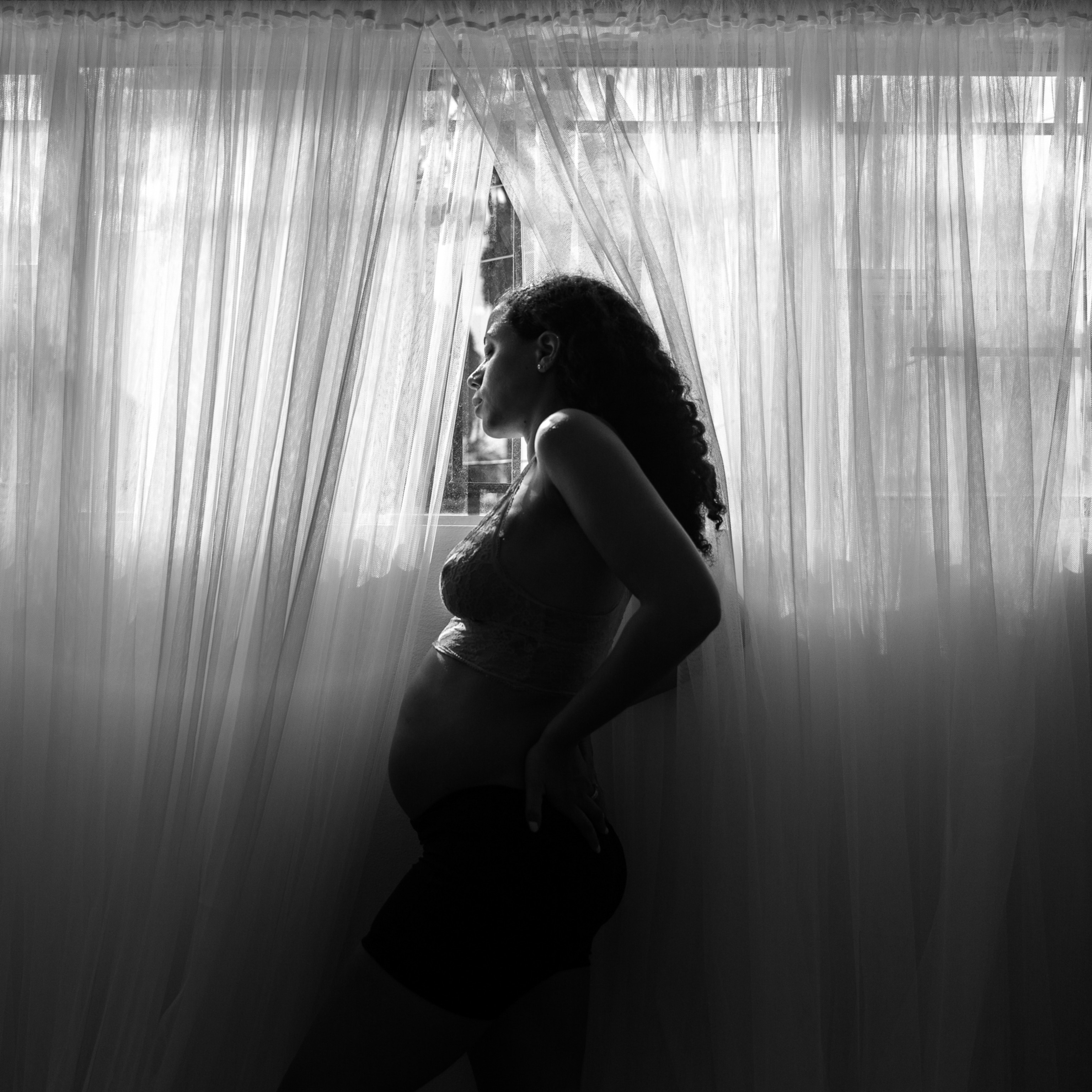 A pregnant woman leans against a window with sheer curtains