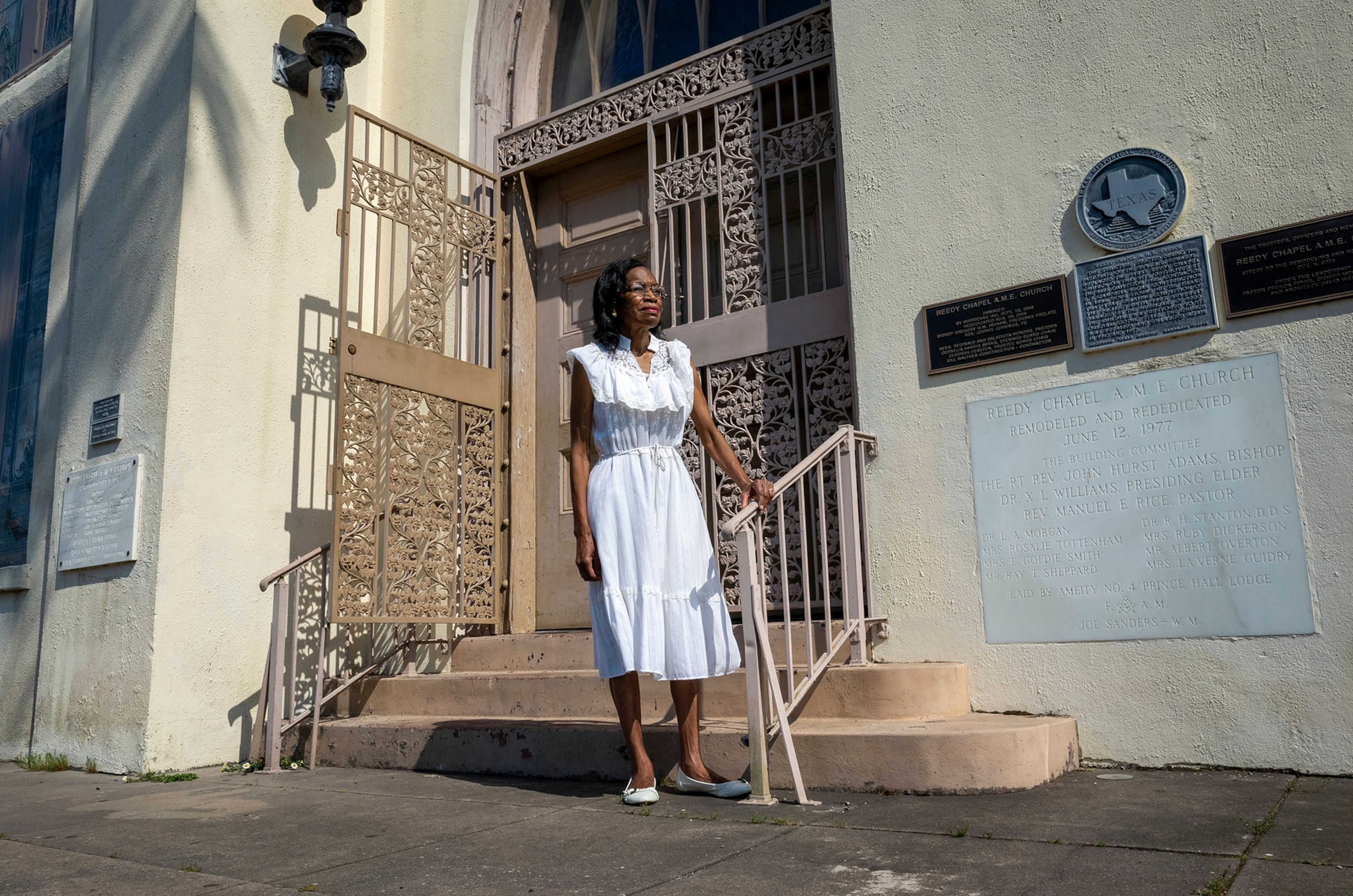 A Black woman wearing a white dress stands in front of a building with a tan iron gate in front of the door.