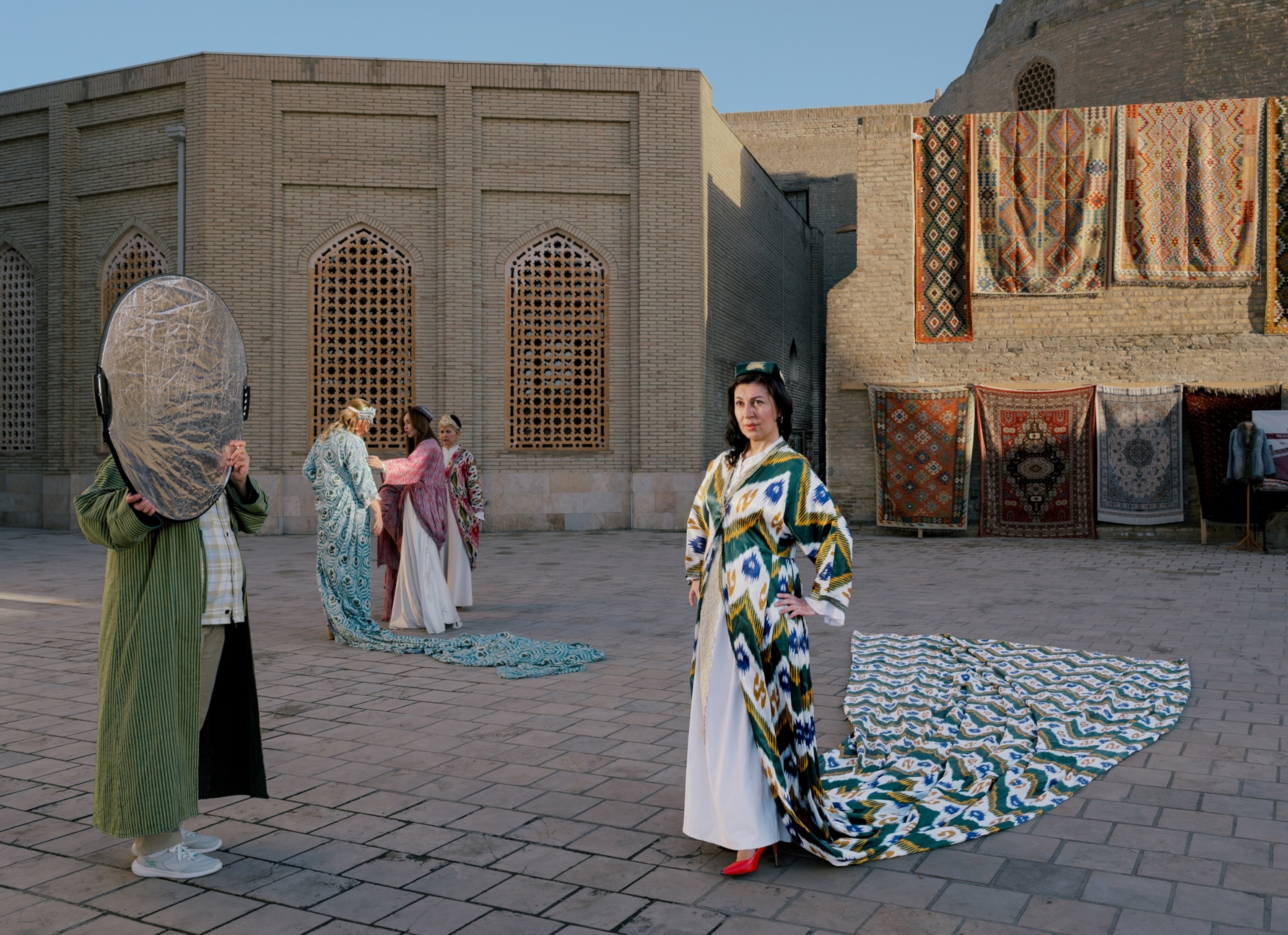 On a tiled city sidewalk, rugs hang in the background on a wall. In the foreground, a woman poses for an image while a person to her left holds up a silver reflector. In the background another group of three poses on the street.