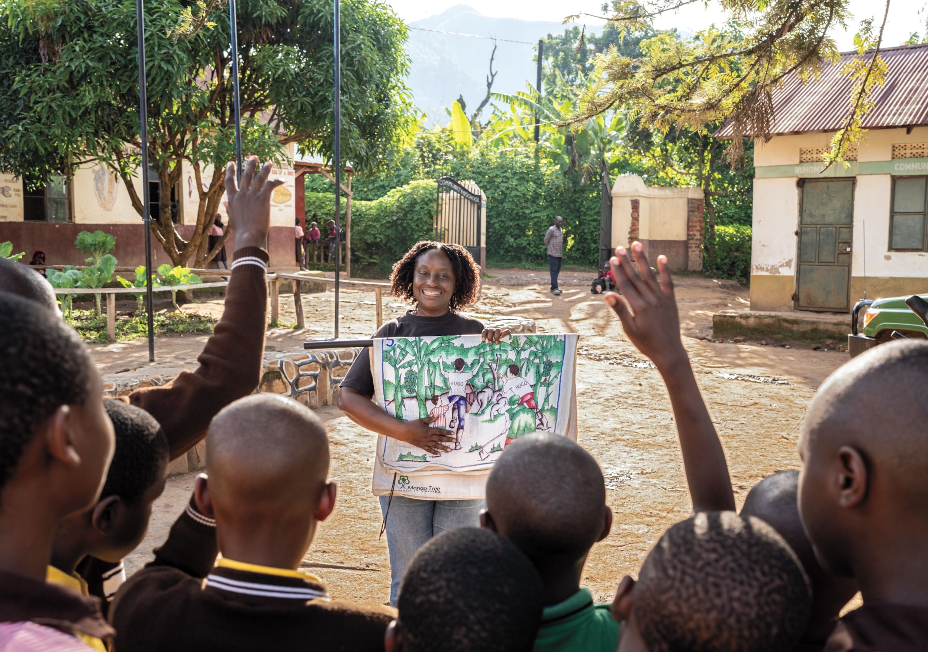 A smiling woman faces the camera and points at a poster with a drawing. In front of her are several school children, and two of them raise their hands.