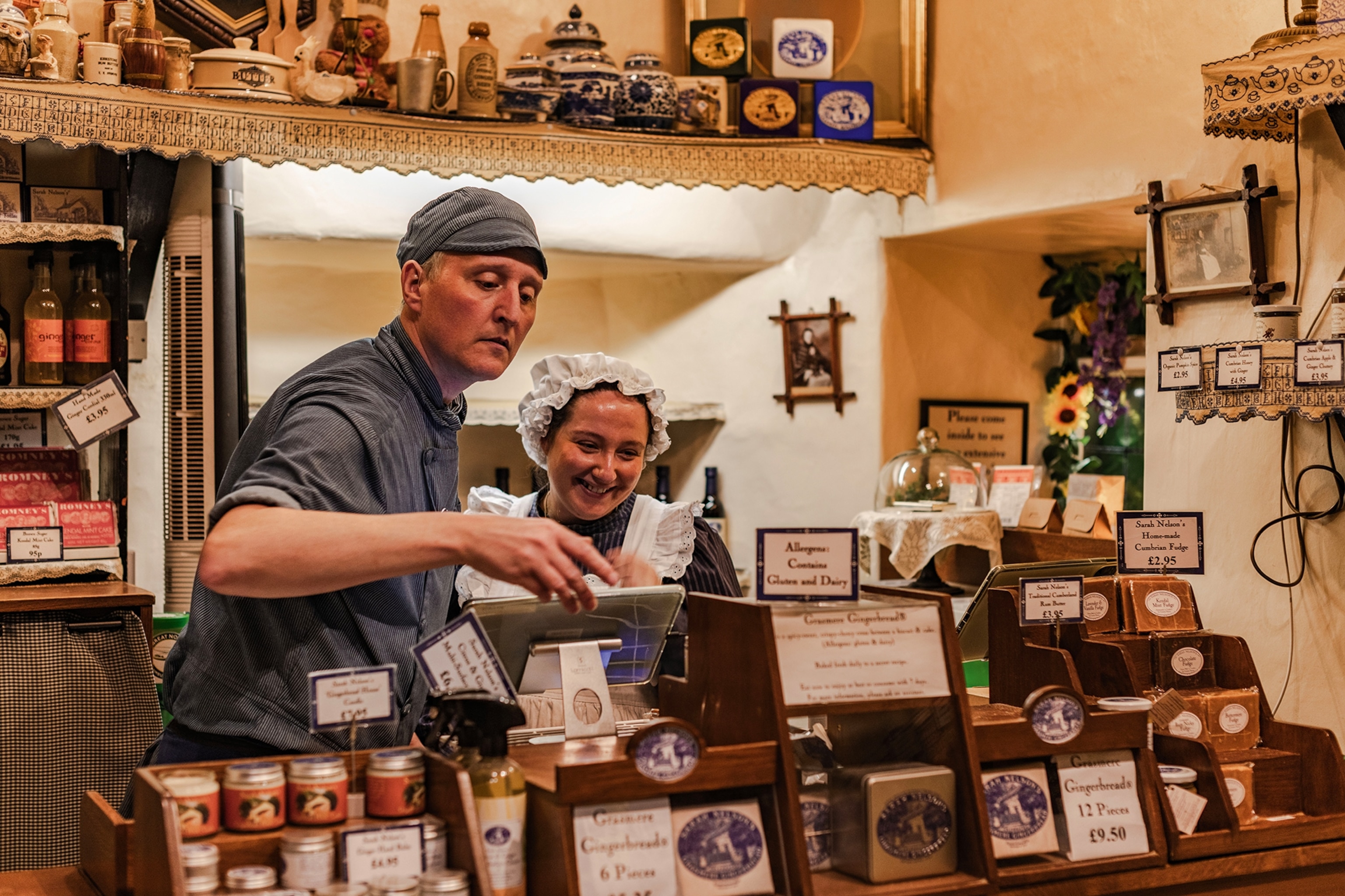 A man and woman dressed in medieval English attire behind a display of gingerbread in a shop.