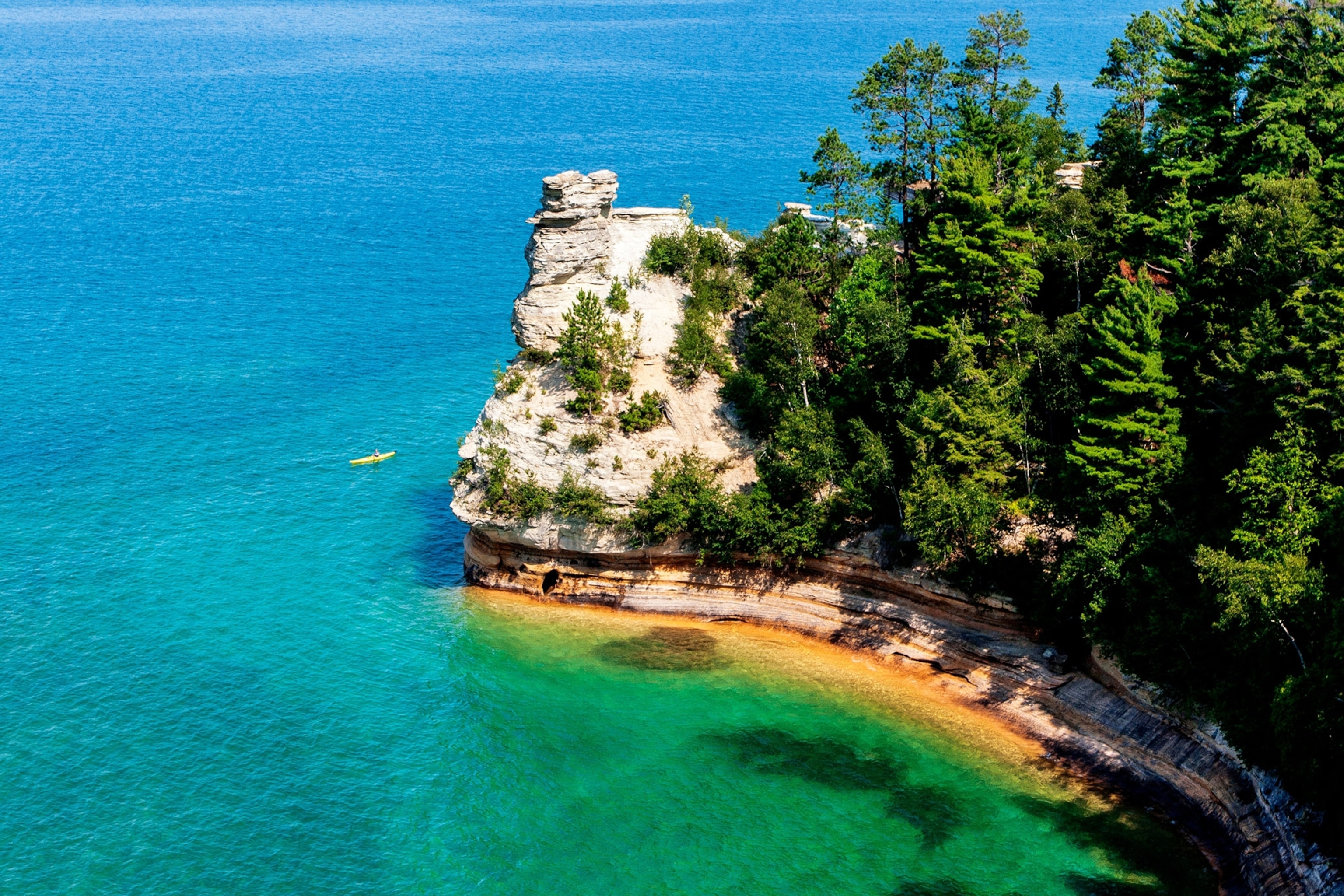 View from above of narrow rocky peninsula topped with trees, surrounded by vibrant blue waters, a kayak passing by
