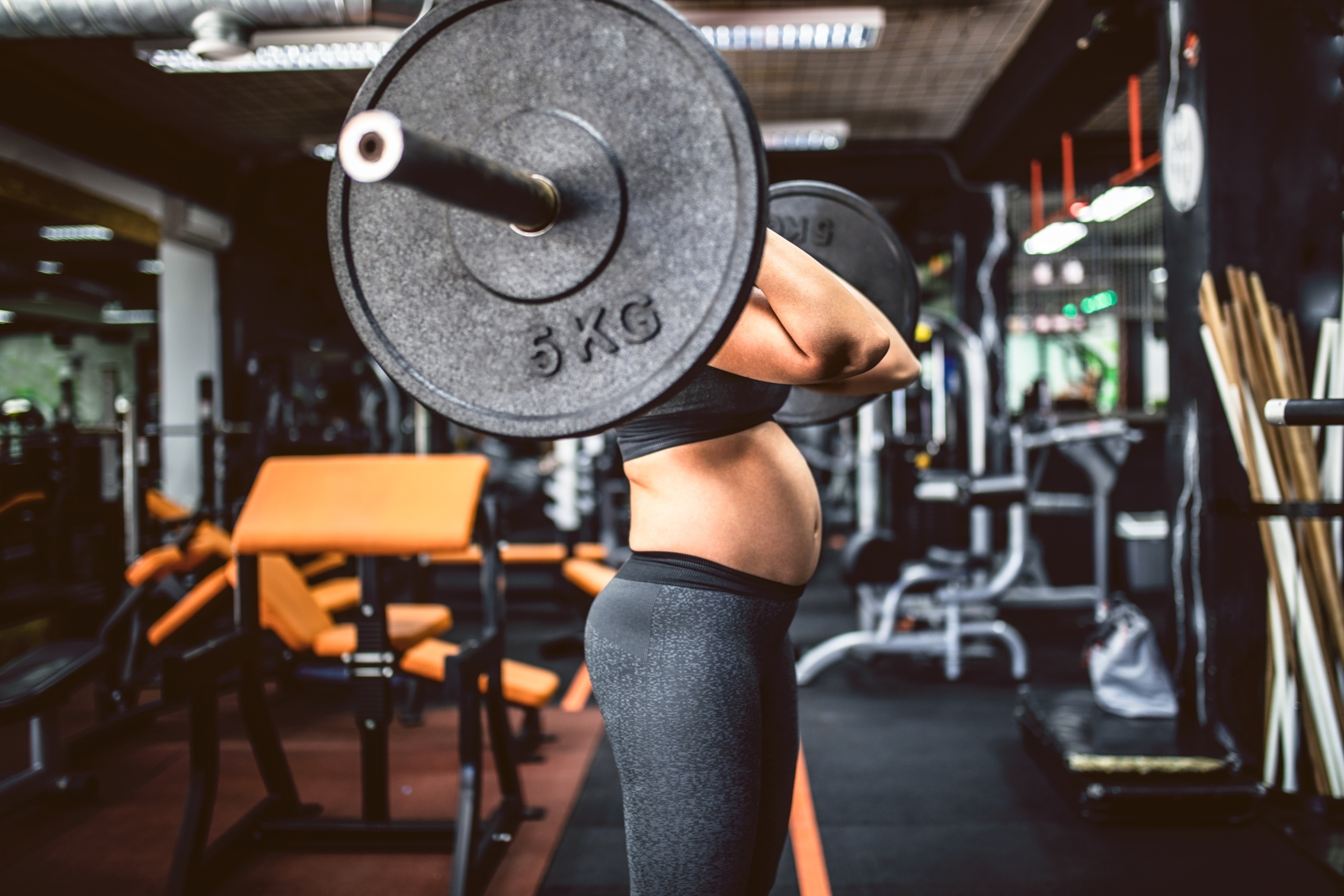 A pregnant woman in workout attire lifting a barbell in a gym. The weight hides her face, drawing attention to her pregnant belly.