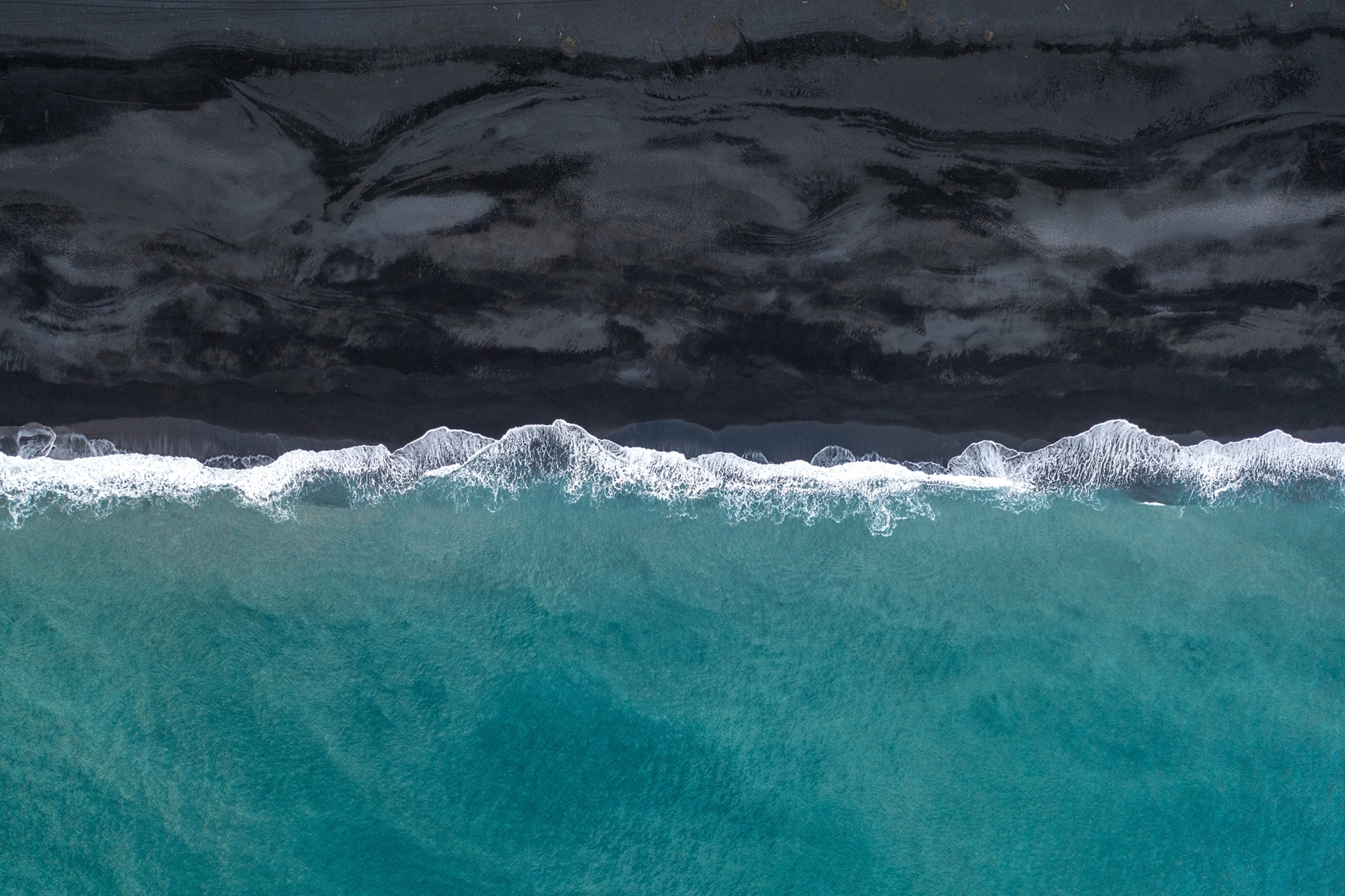 An aerial view over a black sand beach with waves crashing on it.