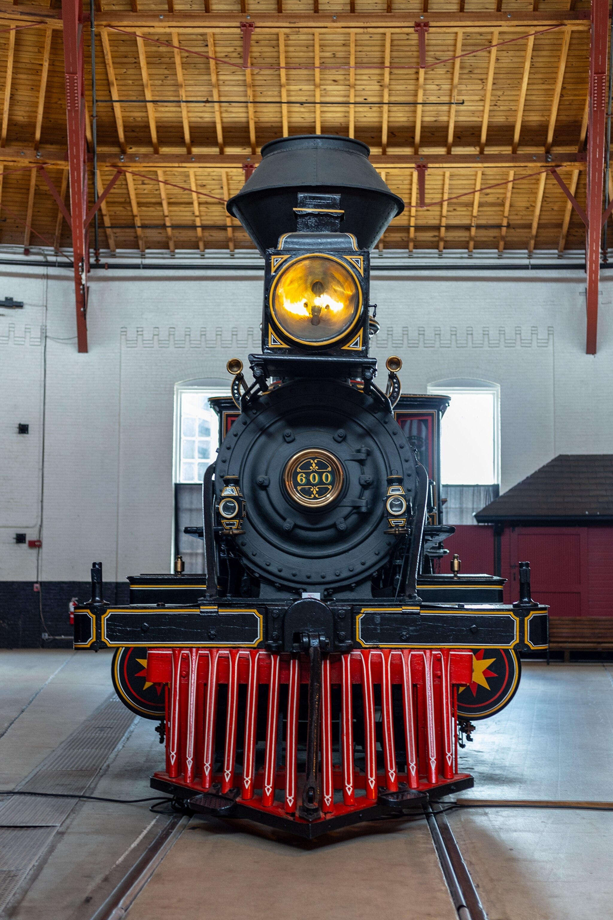 A front view of a historic black steam locomotive with a glowing headlight and bright red cowcatcher, displayed inside a railway museum with high wooden ceilings and train tracks running beneath it.