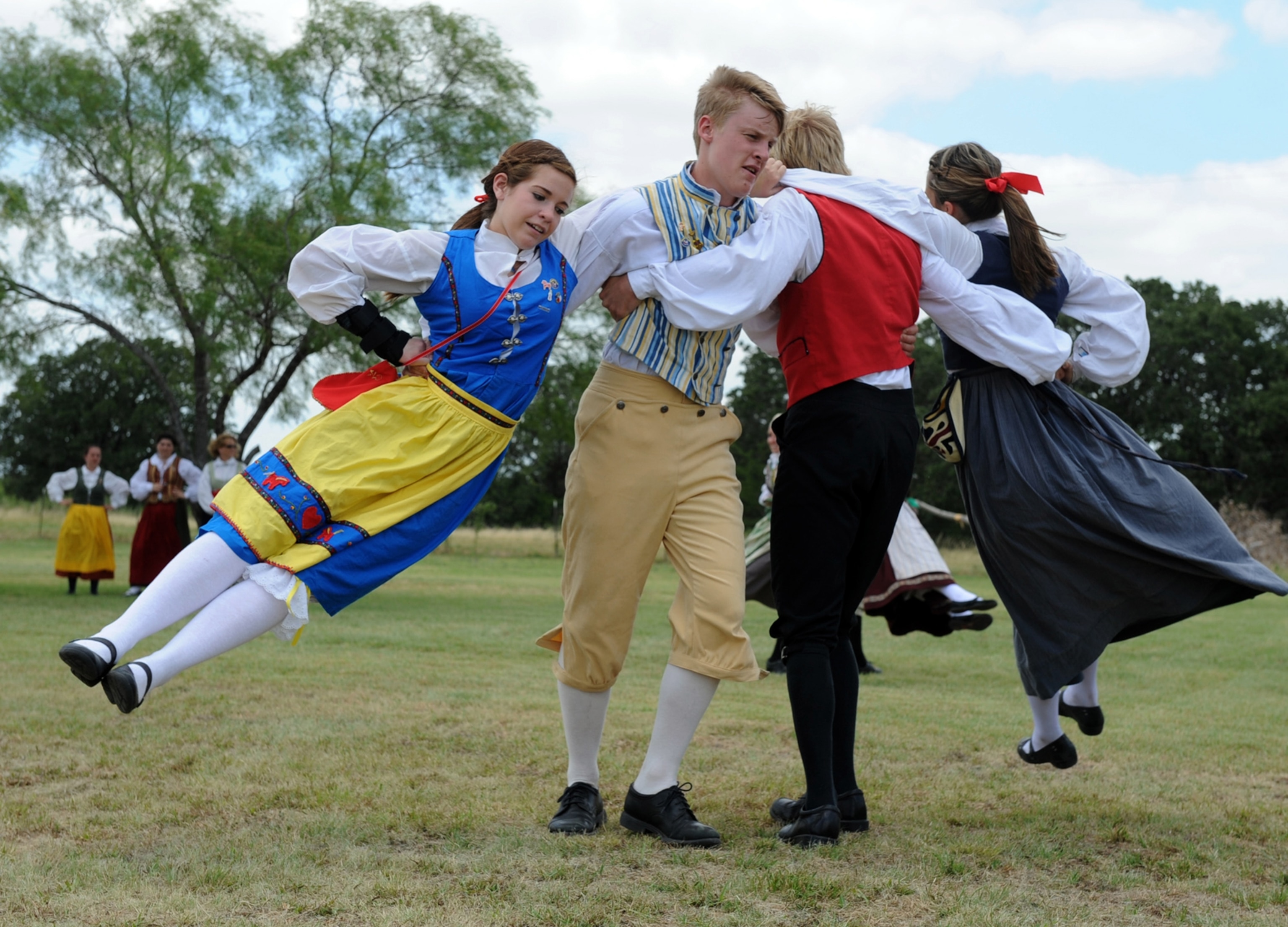 A group of people in colorful traditional costumes dance joyfully in a grassy field
