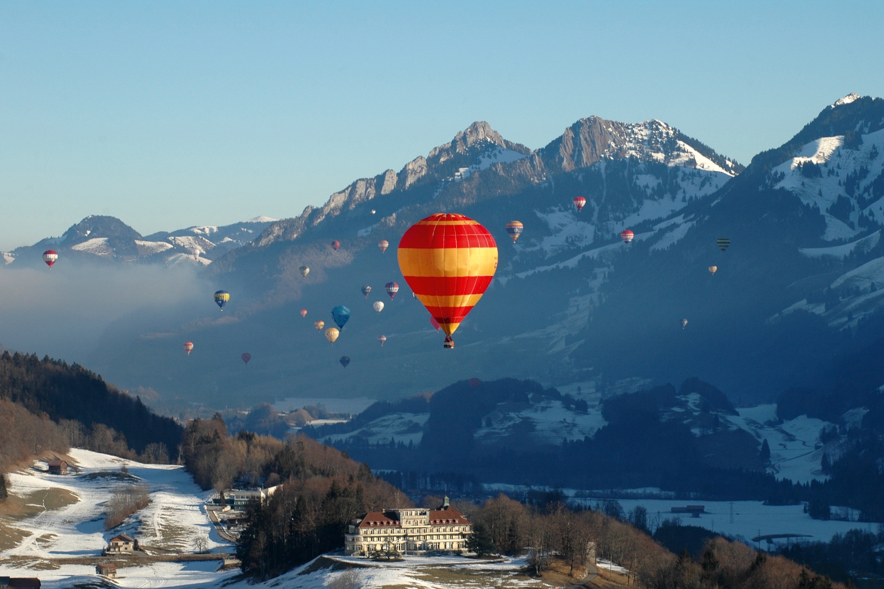 A hot air balloon in the focus as a dozen more float in the sky behind, a snowy mountain scape stretching out across the background.