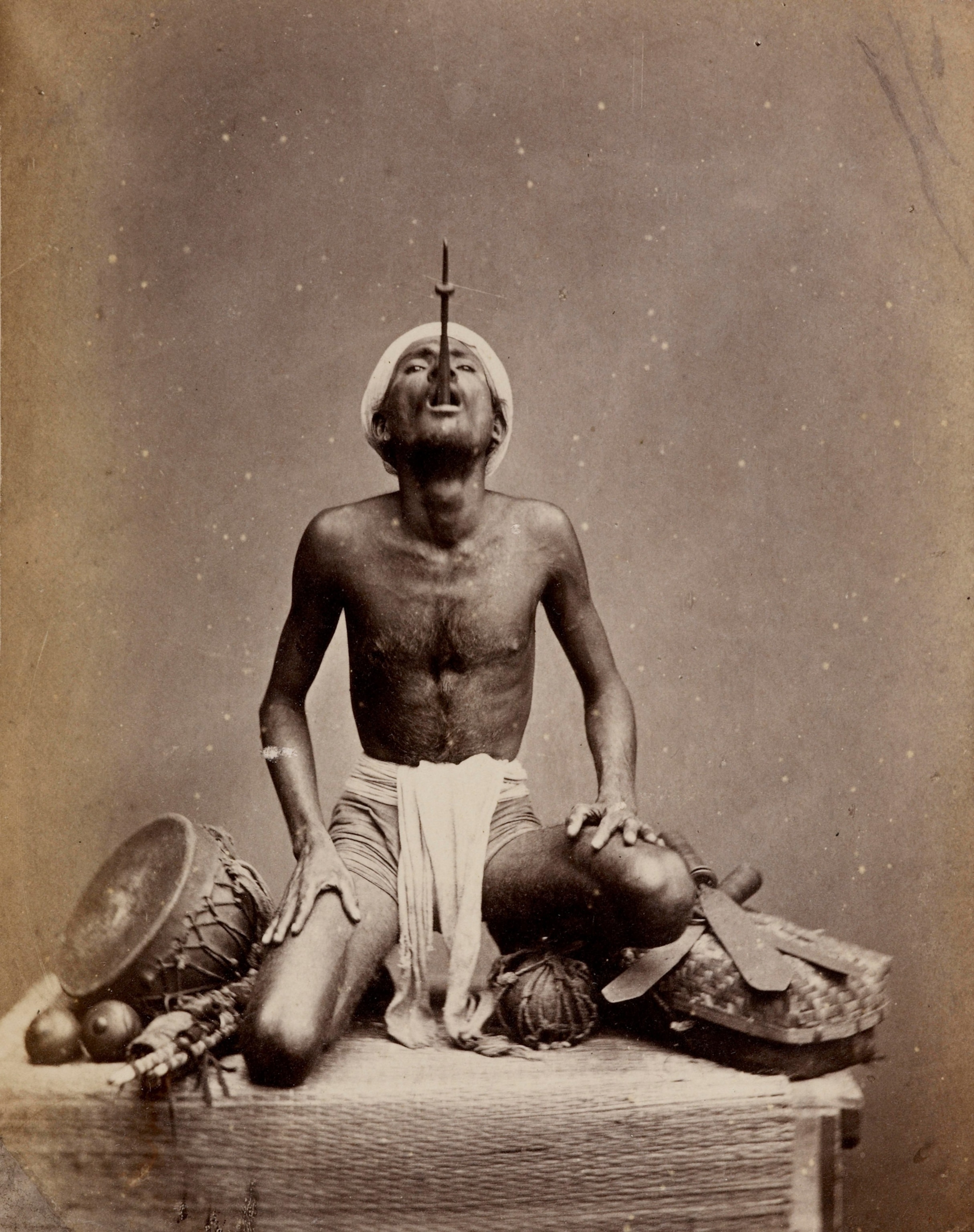 Studio portrait of an Indian juggler who is seated crosslegged, with the hilt of a sword protruding from his mouth