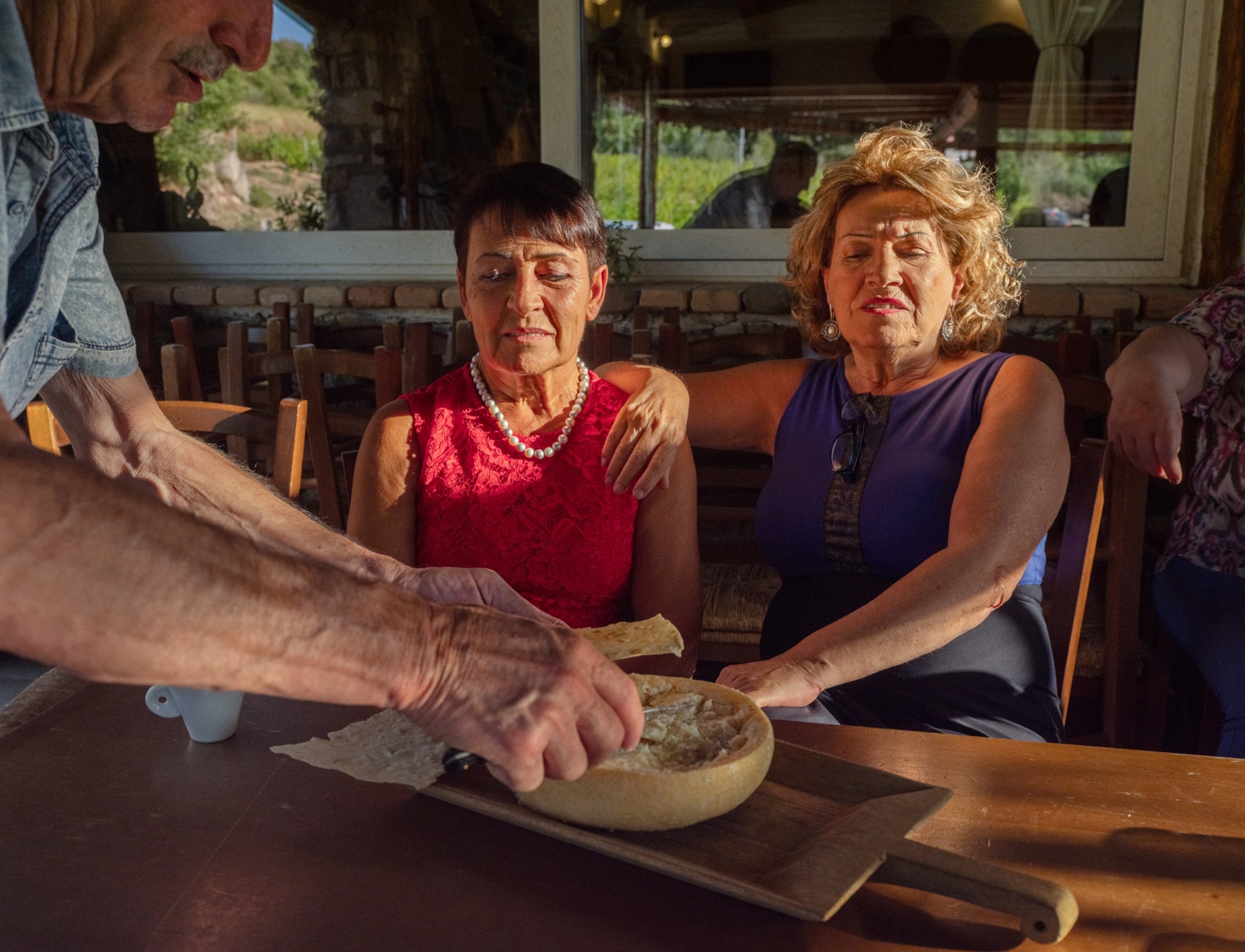 Two women looking at the opened wheel of cheese from which man is scooping interior.