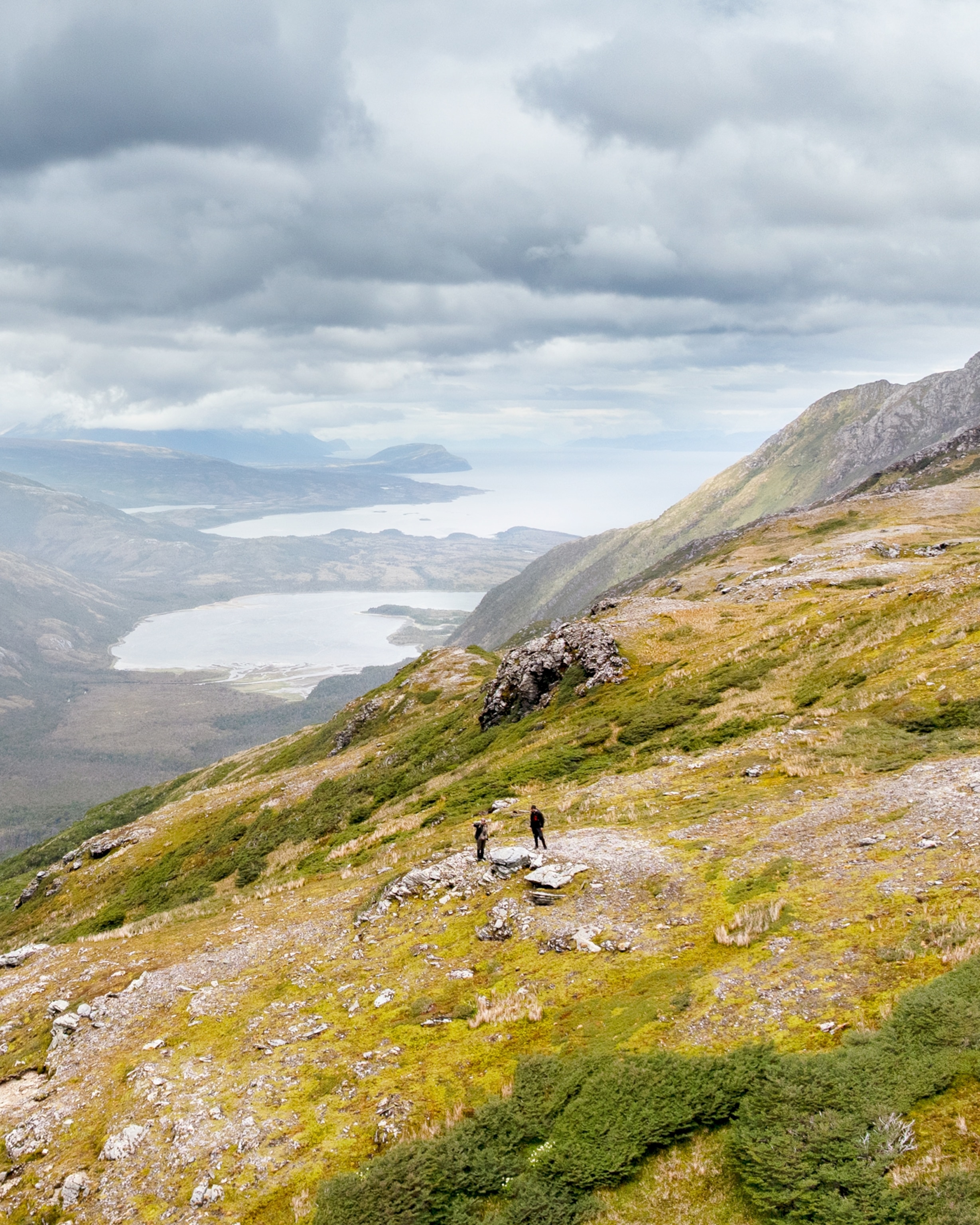 Two hikers stand on a grassy mountain slope overlooking a series of lakes and valleys, with rugged peaks and dark clouds stretching across the landscape in the distance.