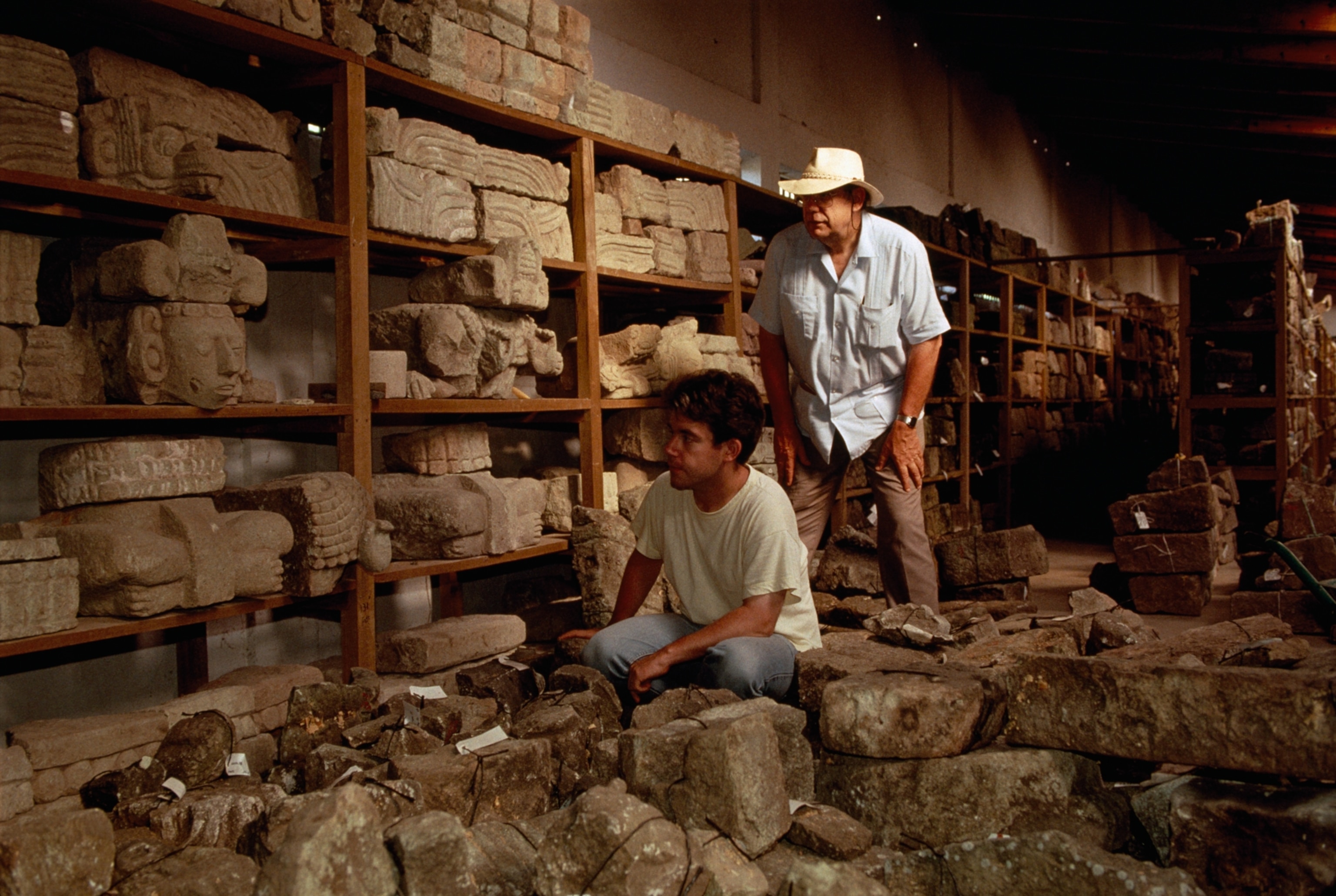 Two men in an archaeological storage room examine ancient stone carvings, surrounded by shelves filled with artifacts