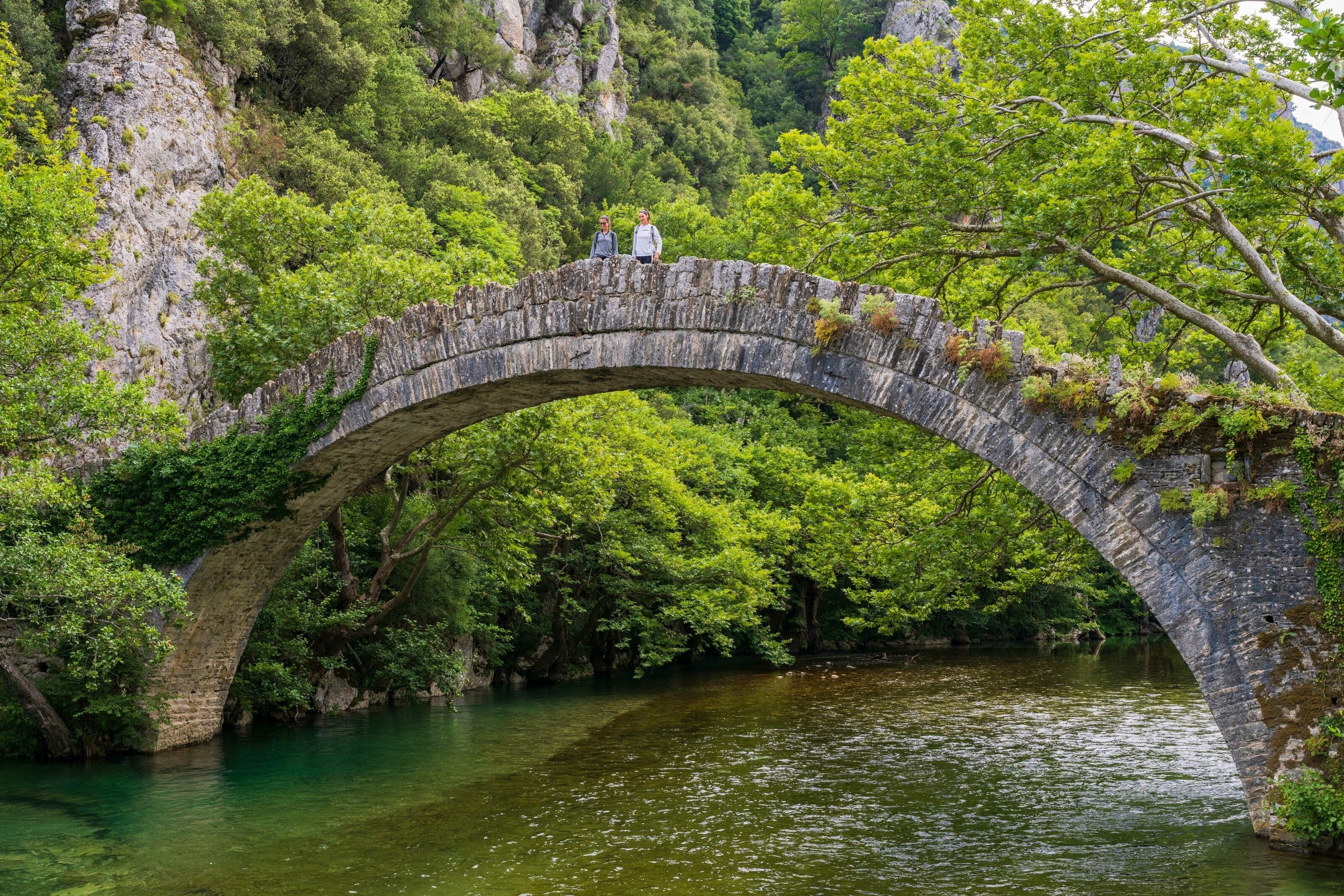 The Kleidonia bridge is made of stone, it passes over a glassy river between two green banks with large and vibrant trees.