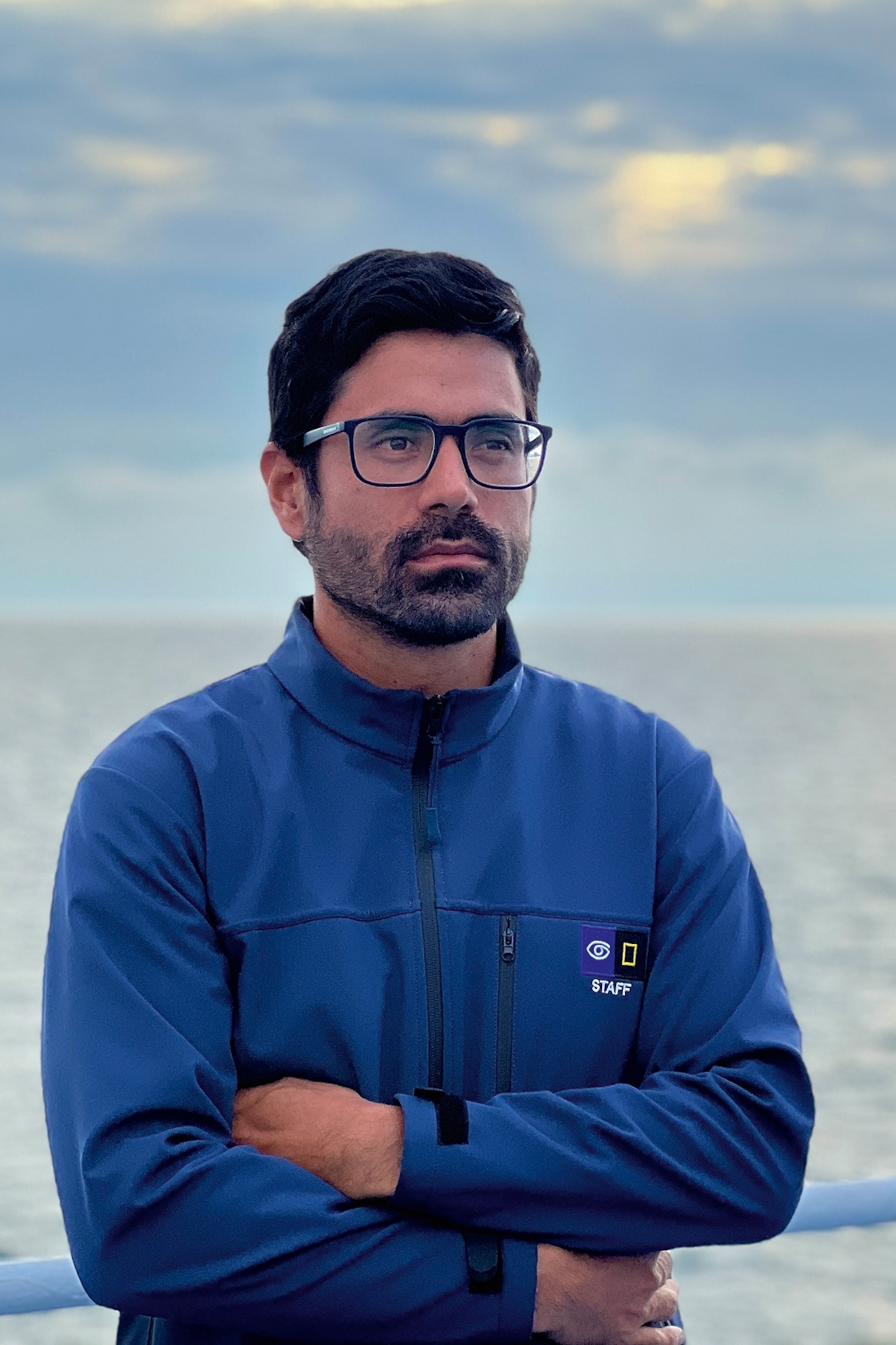 A stern-looking local with slight beard on a ship, wearing glasses and a windbreaker.
