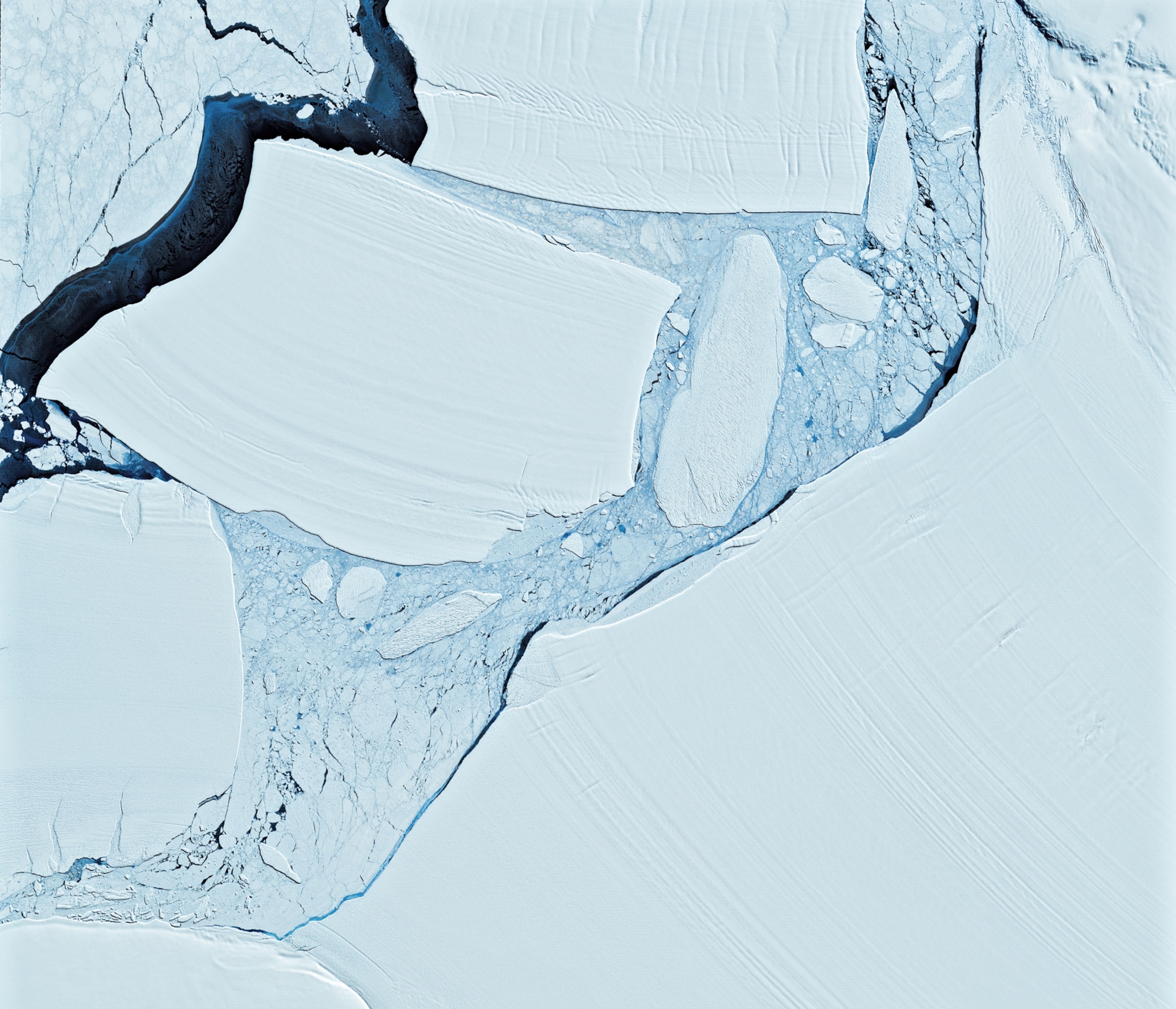 Aerial view of cracked ice sheets with smooth and jagged edges, separated by dark crevices.