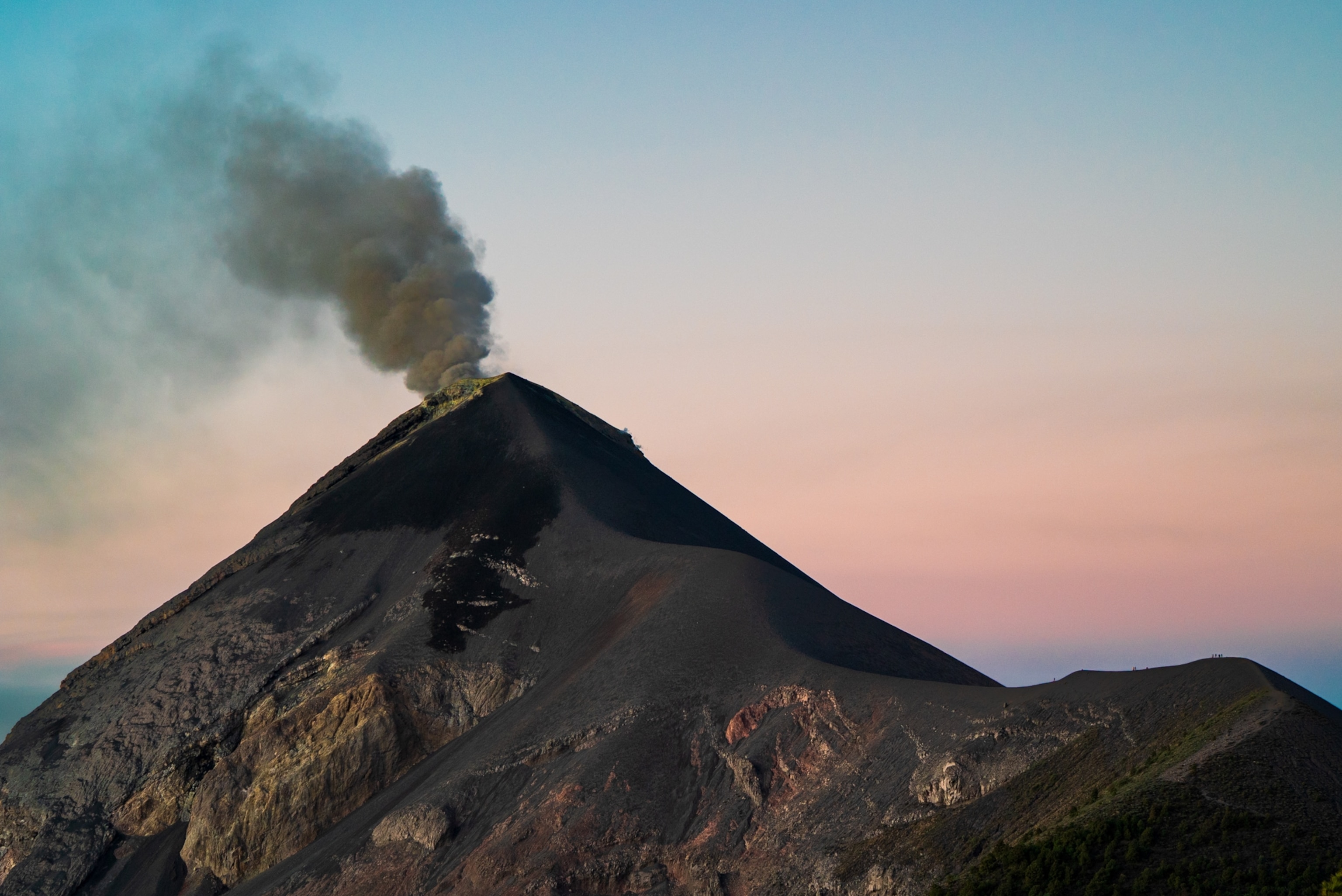 A dark volcanic peak emits thick smoke against a serene sunset sky with pink and blue hues.