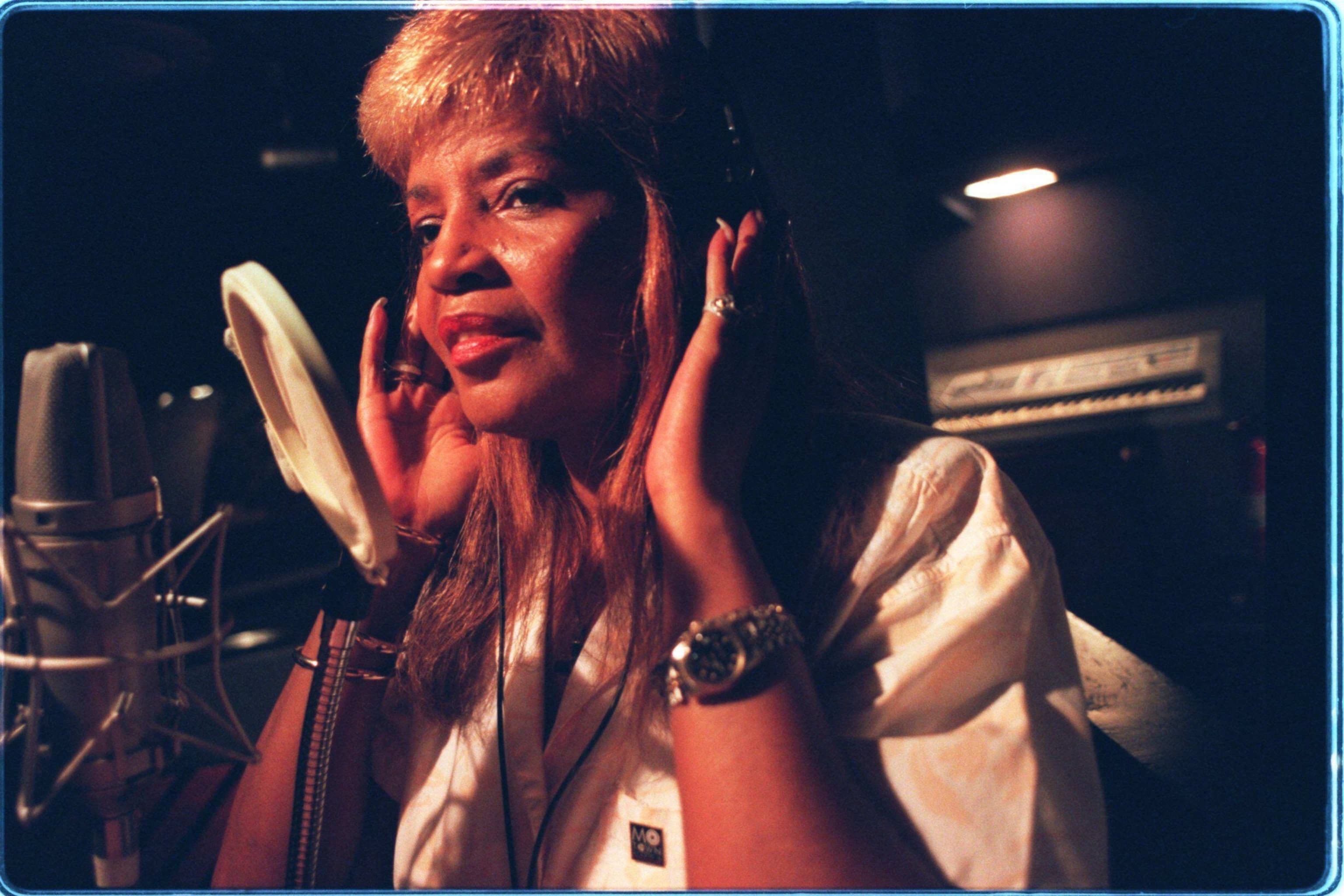 A woman in a recording studio wearing headphones and a white shirt, focused and singing into a microphone