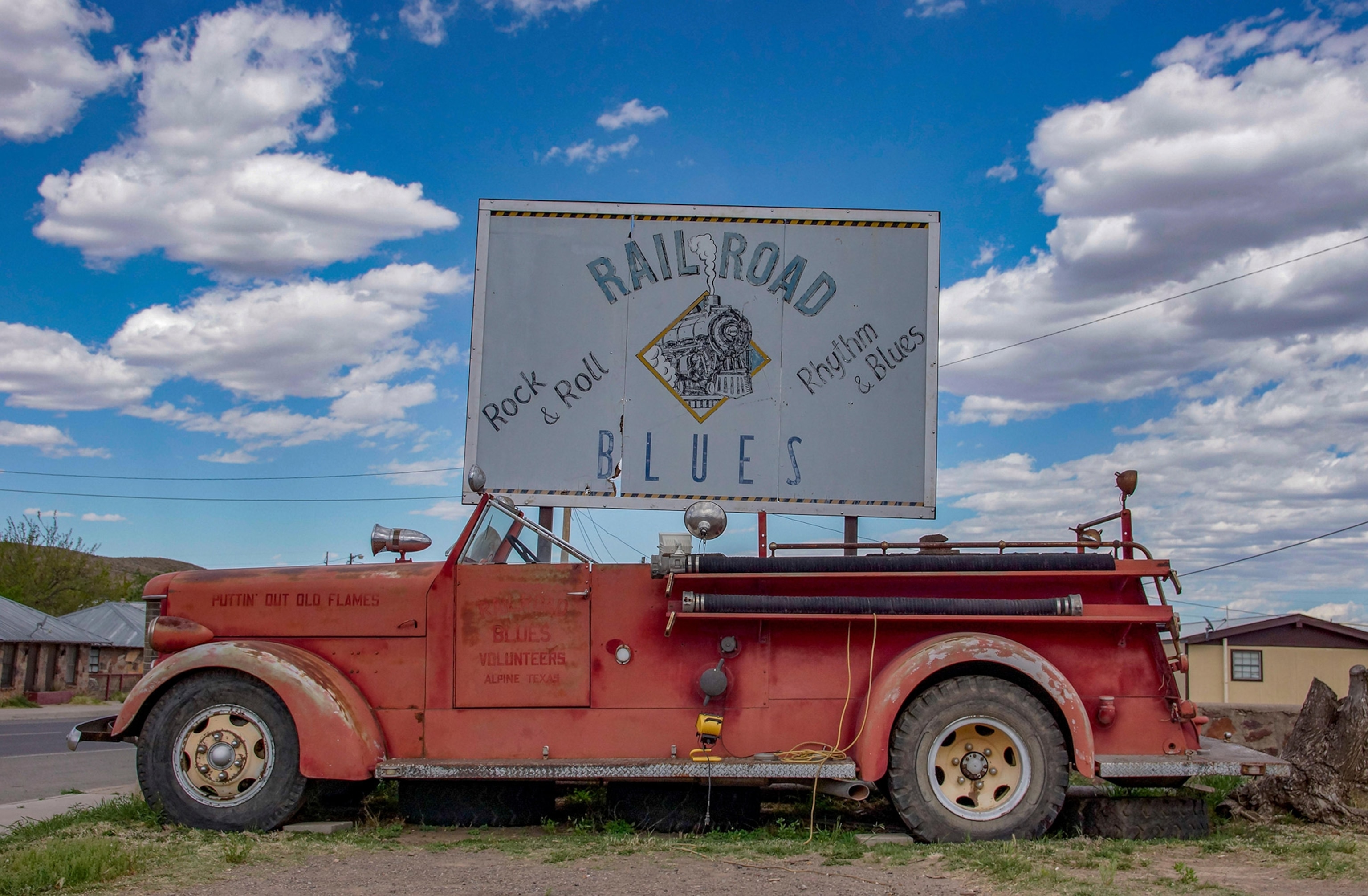 An old rusty red truck stands on a roadside, a sign propped up above it reading "Railroad Blues", blue sky with puffy clouds