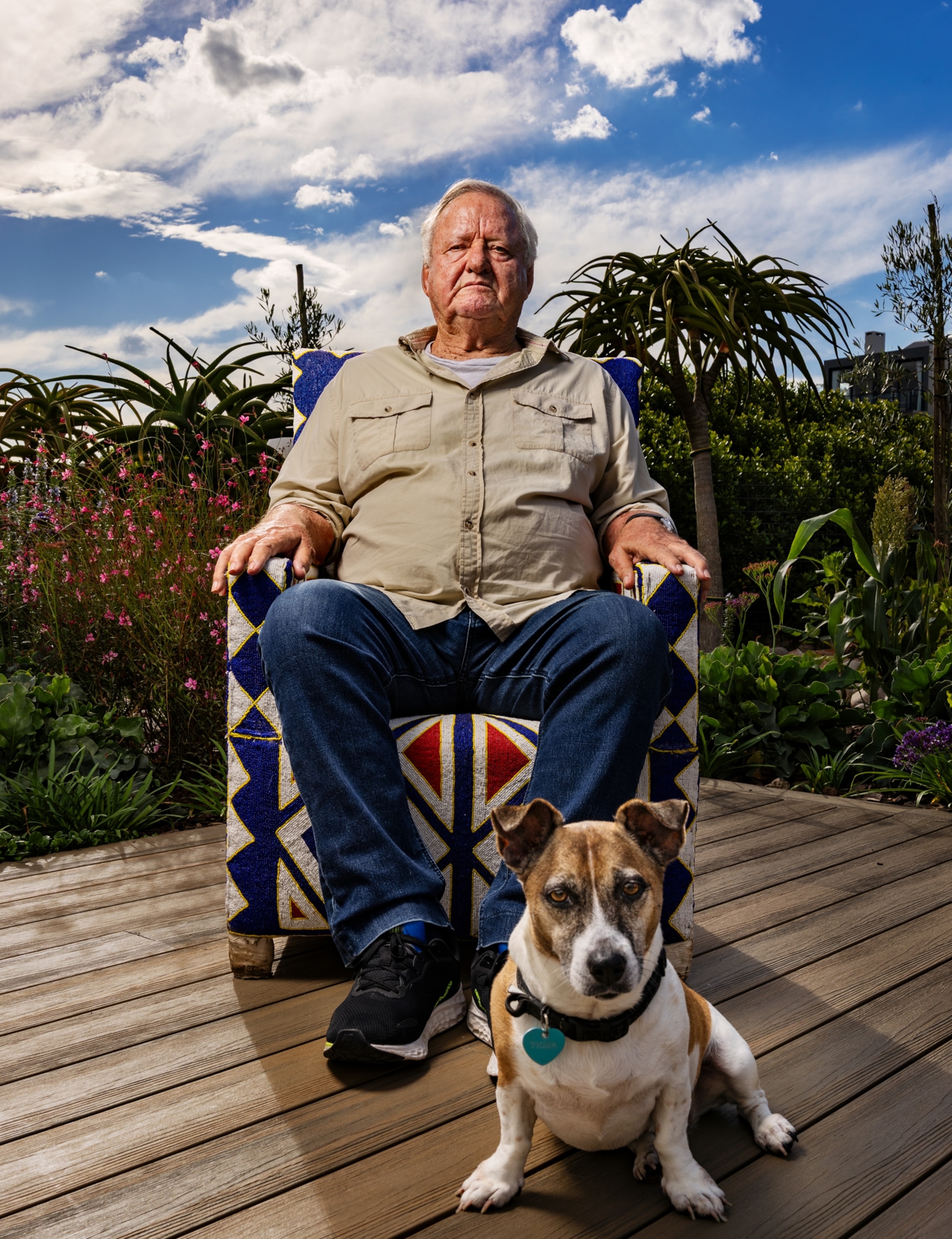 An older man wearing a tan button up and blue jeans sits in a chair on a patio outside. In front of him is a brown and white dog, and they both look directly into the camera as his portrait is taken.