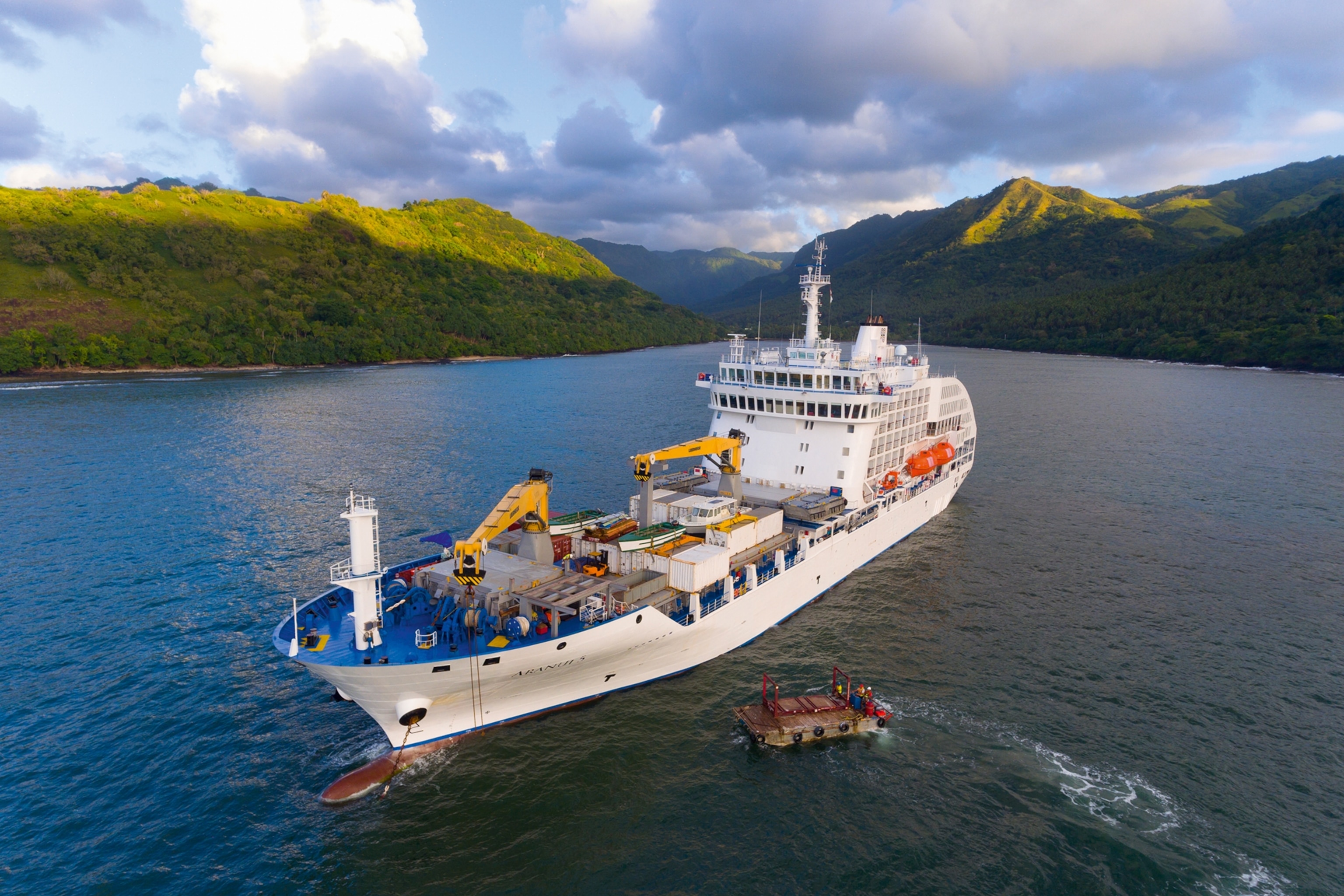 An aerial shot of a cargo cruise ship heading towards a lush island.