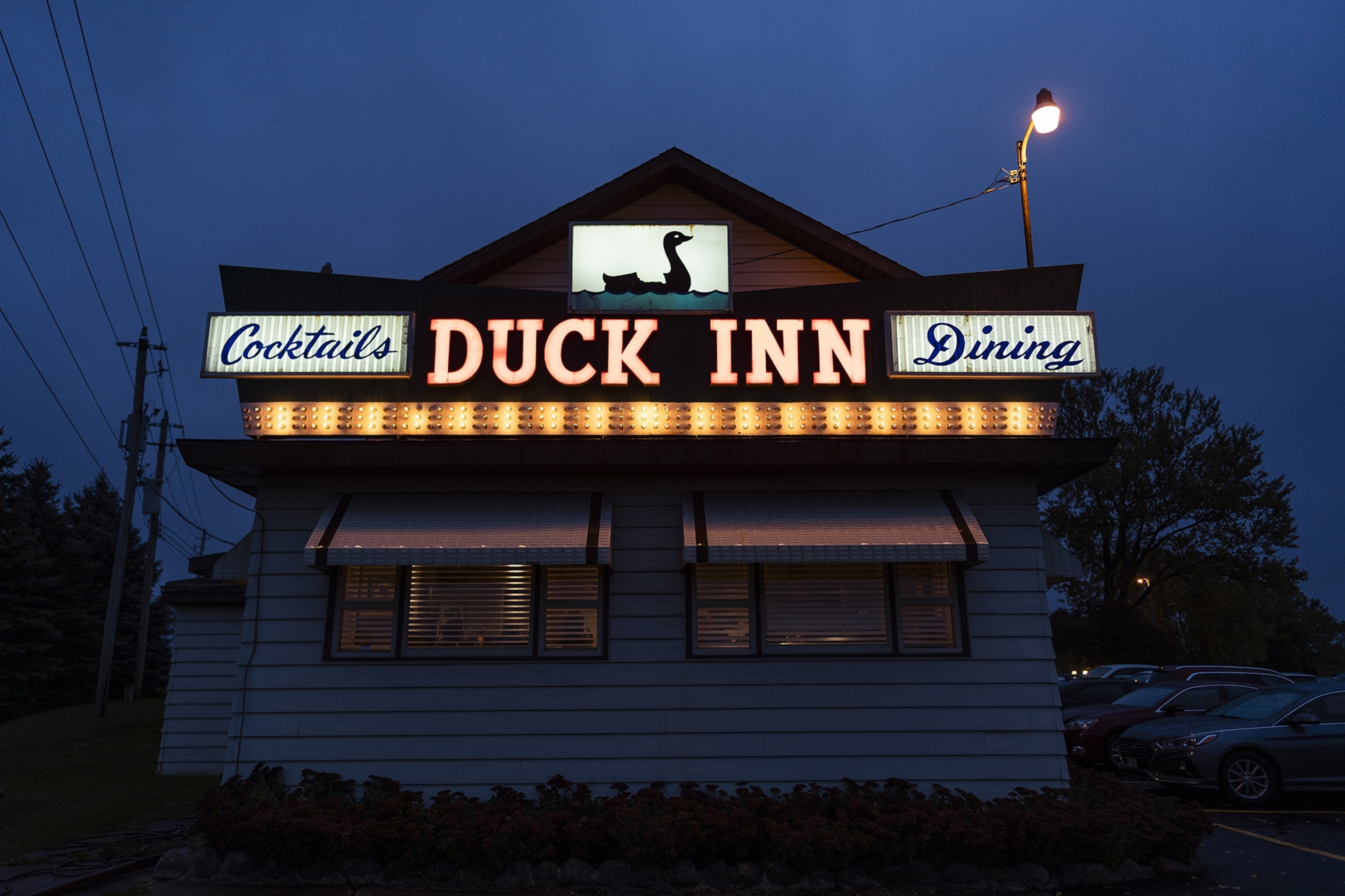 A nighttime exterior of a small building with a glowing sign reading "DUCK INN" with "Cocktails" and "Dining" with a graphic of a duck