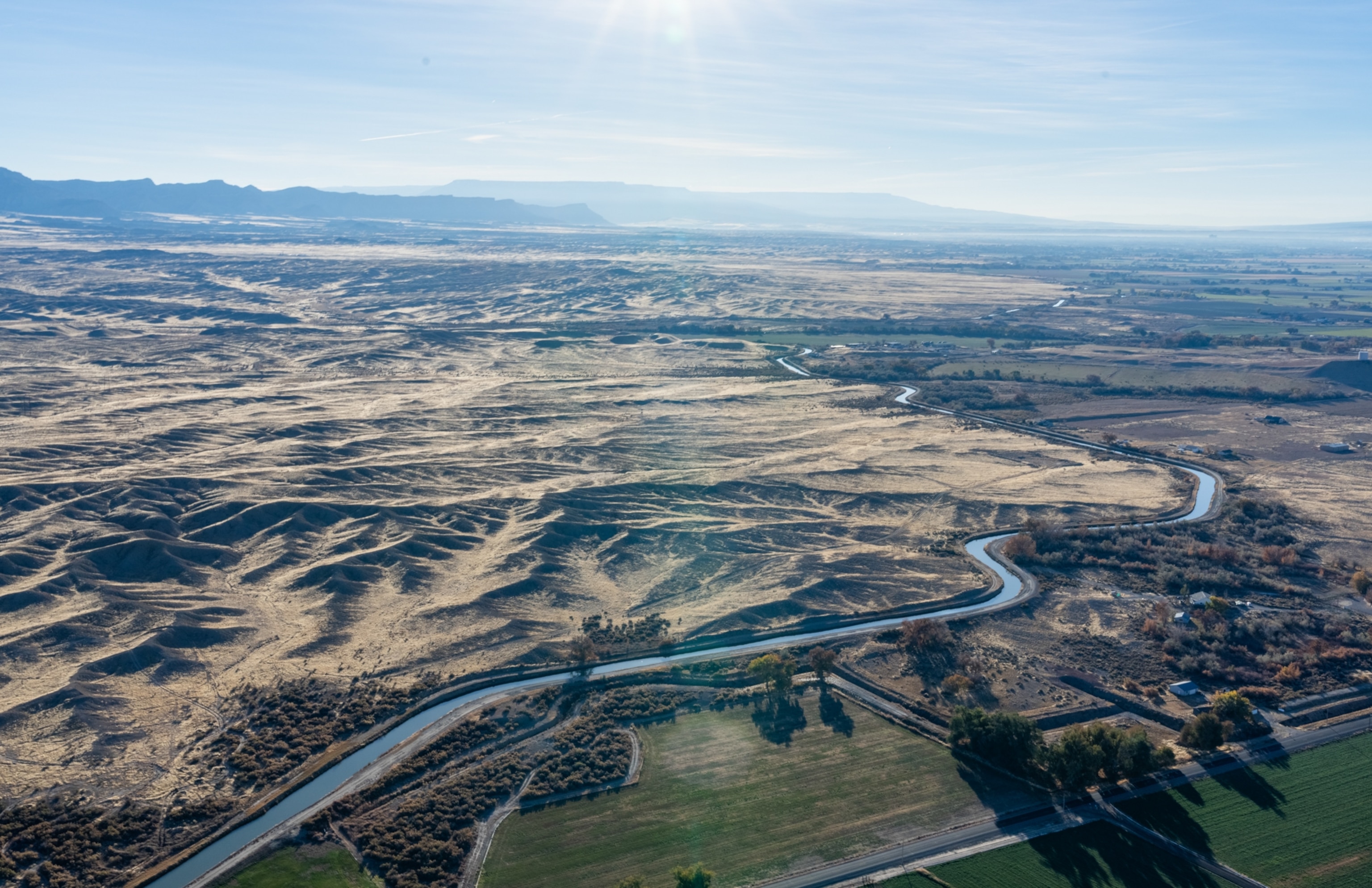 Aerial view of a vast, sunlit landscape with barren hills on the far side of a thin winding river, and some green fields on the near side of the river